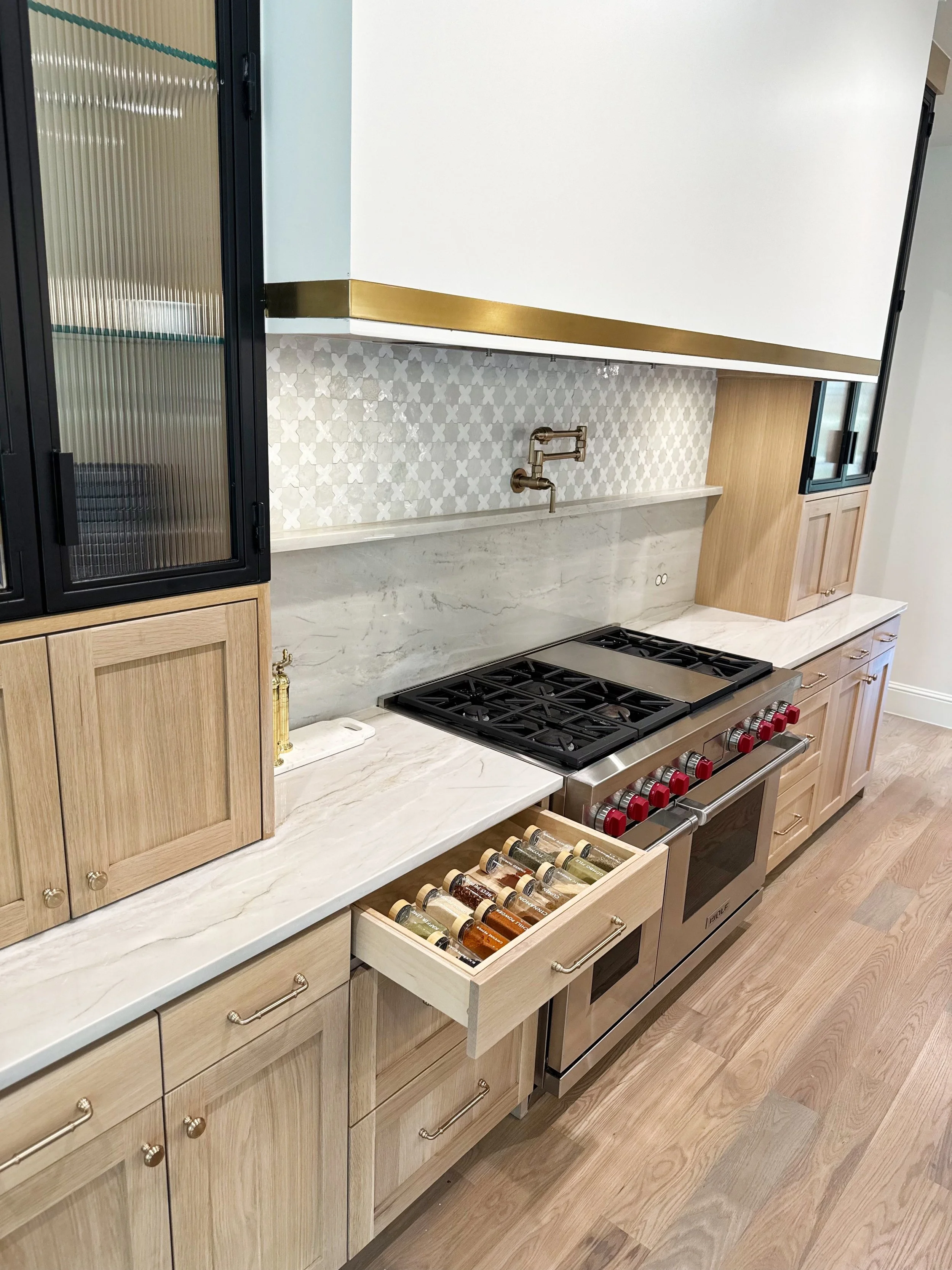 Kitchen with light wood cabinets, a marble countertop, a gas stove with red knobs, and a drawer containing spices. There are glass-front cabinets, a brass faucet, and a decorative tile backsplash.