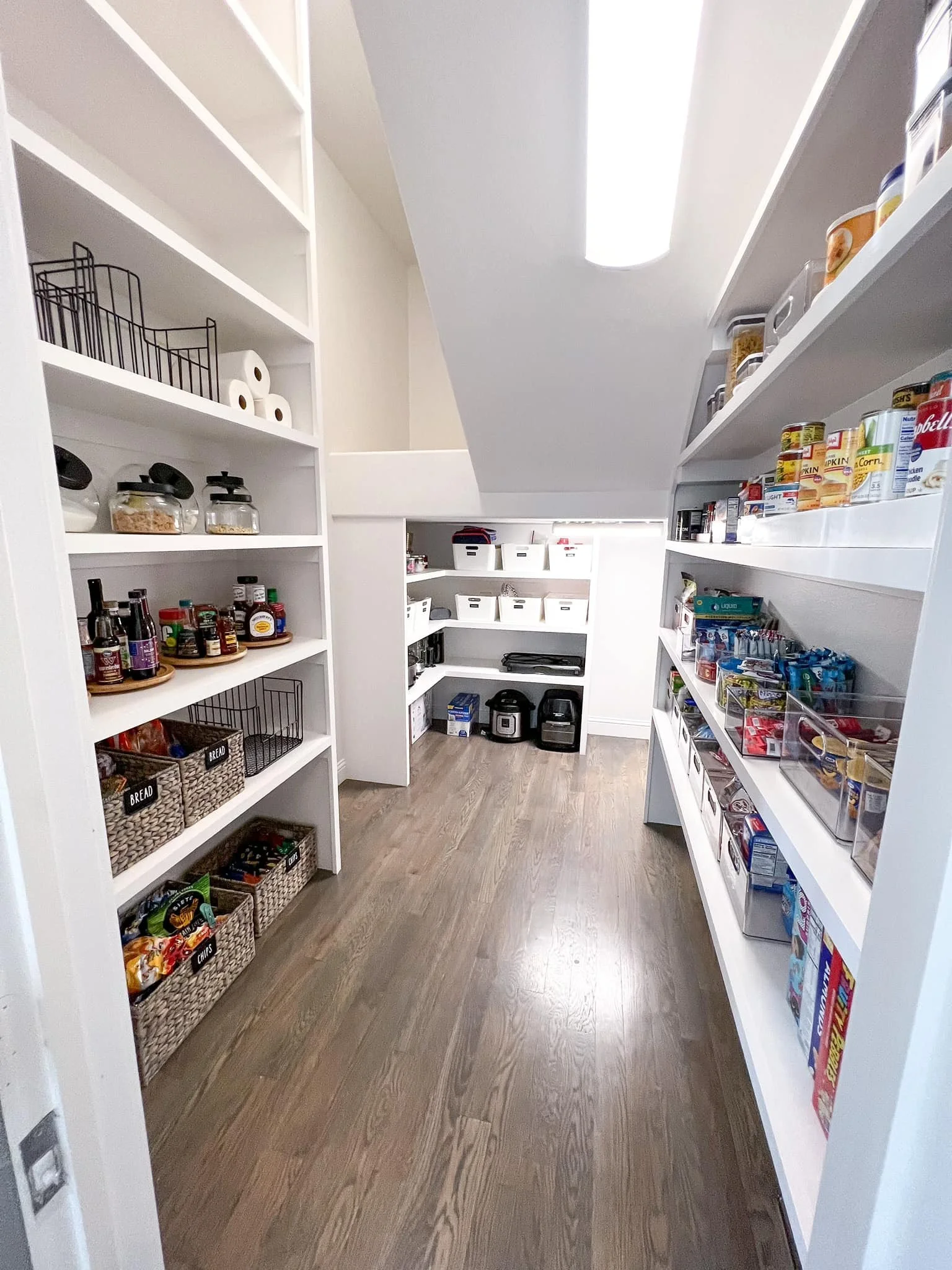 Pantry with white shelves stocked with canned goods, snacks, and condiments, and a wooden floor.