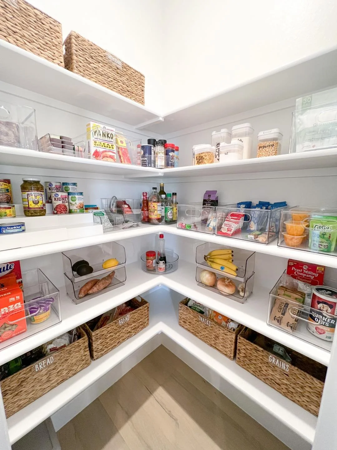 Organized pantry with three white shelves containing various food items in clear containers and baskets, including bread, snacks, grains, bananas, onions, and condiments.