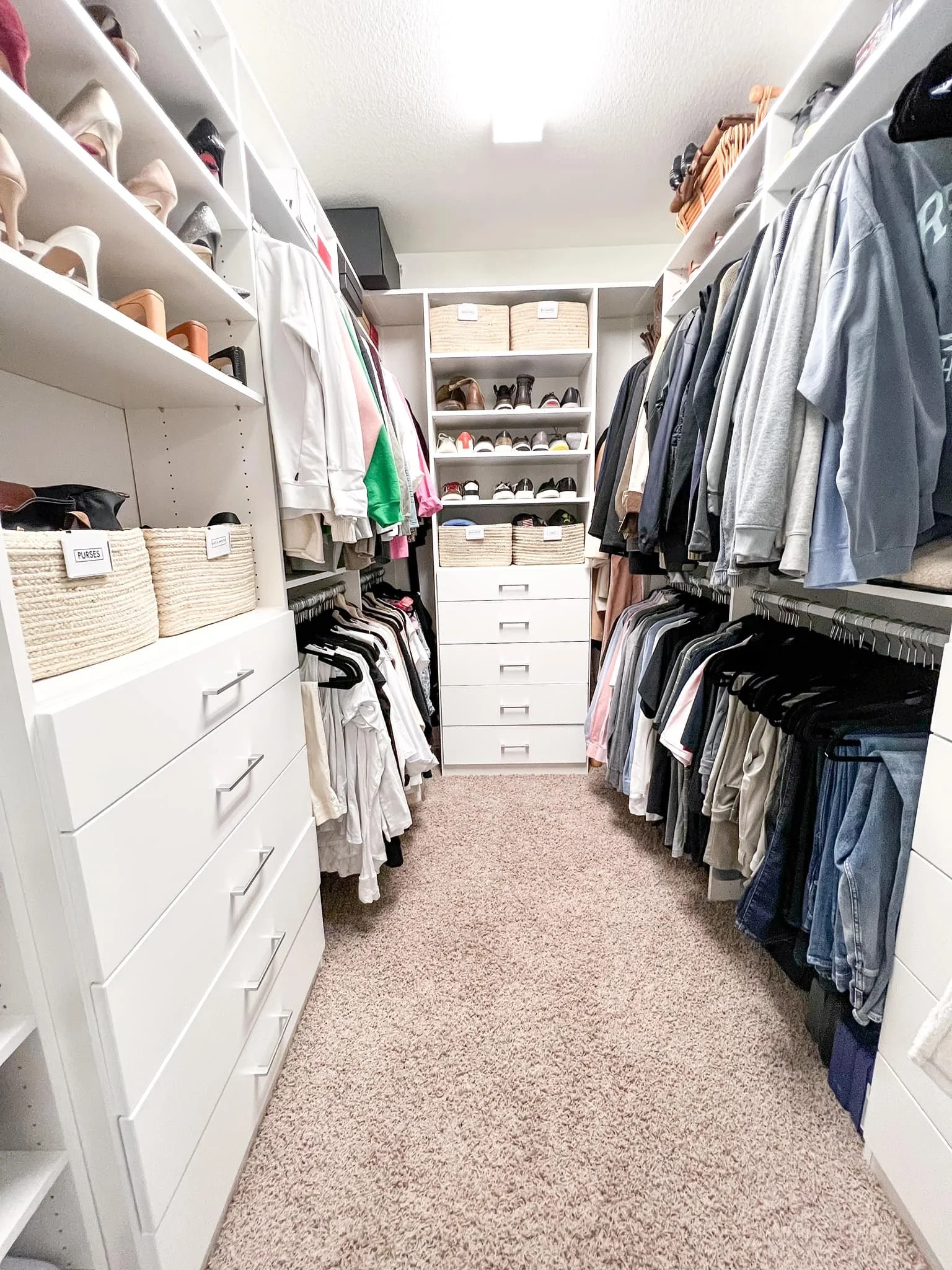 A walk-in closet with white shelving and drawers, filled with shoes, handbags, and clothing. The floor is covered with beige carpet.