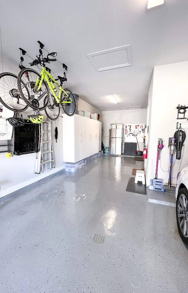 Clean garage with bikes hung on the wall, a ladder, white cabinets, a refrigerator, and cleaning tools near a car.