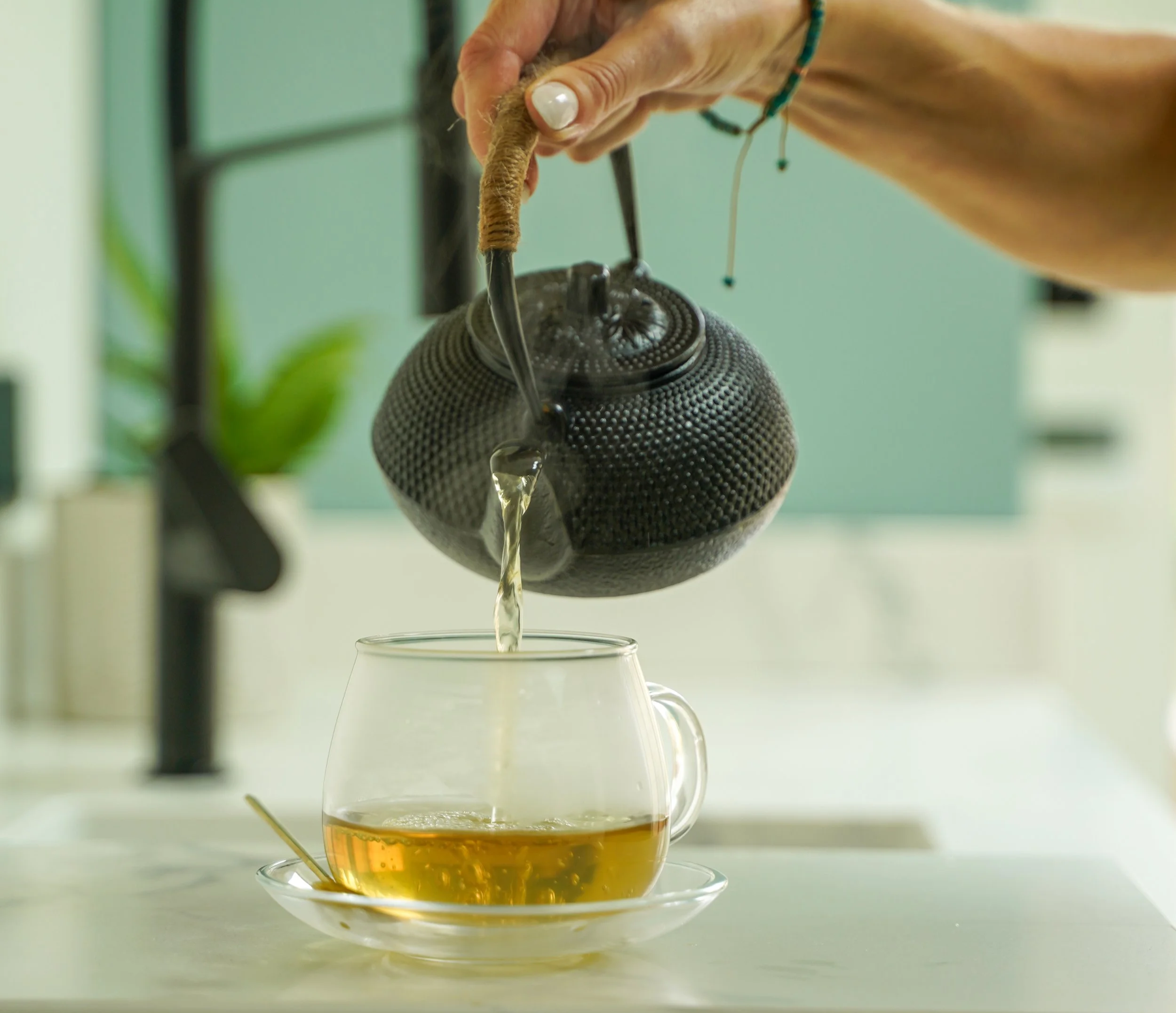 Close-up of a hand pouring an Ayurvedic herbal rose tea from a teapot into a glass cup, with a light green wall and a plant in the background.