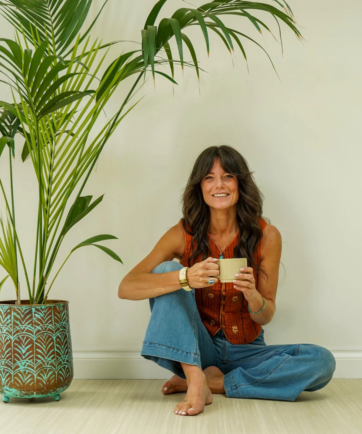 A woman with long dark hair, wearing an orange vintage 70's Indian top and blue jeans, sitting cross-legged & bare foot on the floor. She is smiling and holding a mug of turmeric milk. With a big leaved large potted palm plant next to her.