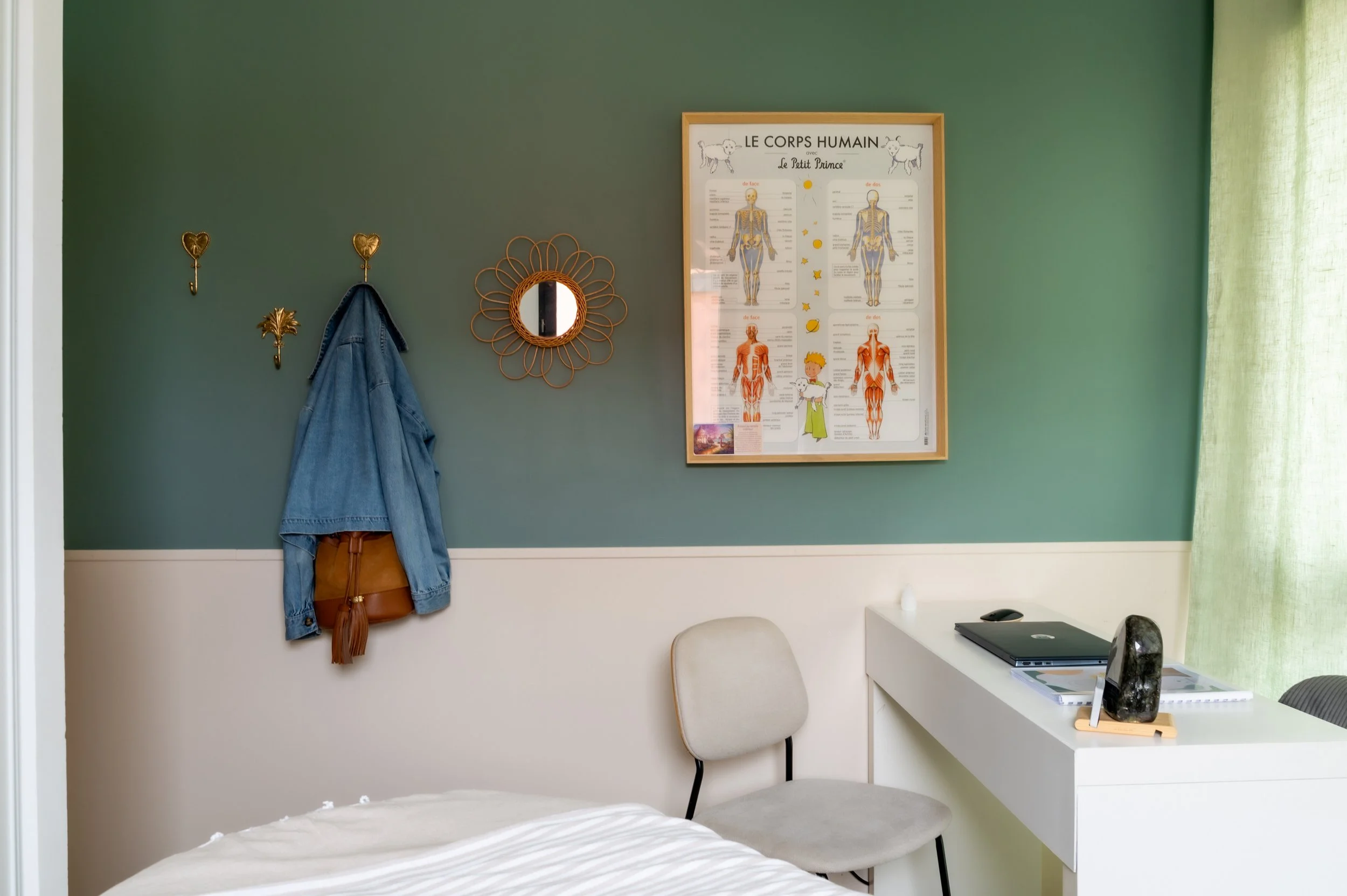 Coin d'une chambre avec un mur vert, un tableau anatomique du corps humain, un miroir en forme de fleur, des crochets décoratifs avec un sac et une veste, un bureau blanc avec un ordinateur portable, une lampe, un carnet et une pierre décorative, une chaise beige et une partie du lit avec une couverture blanche.