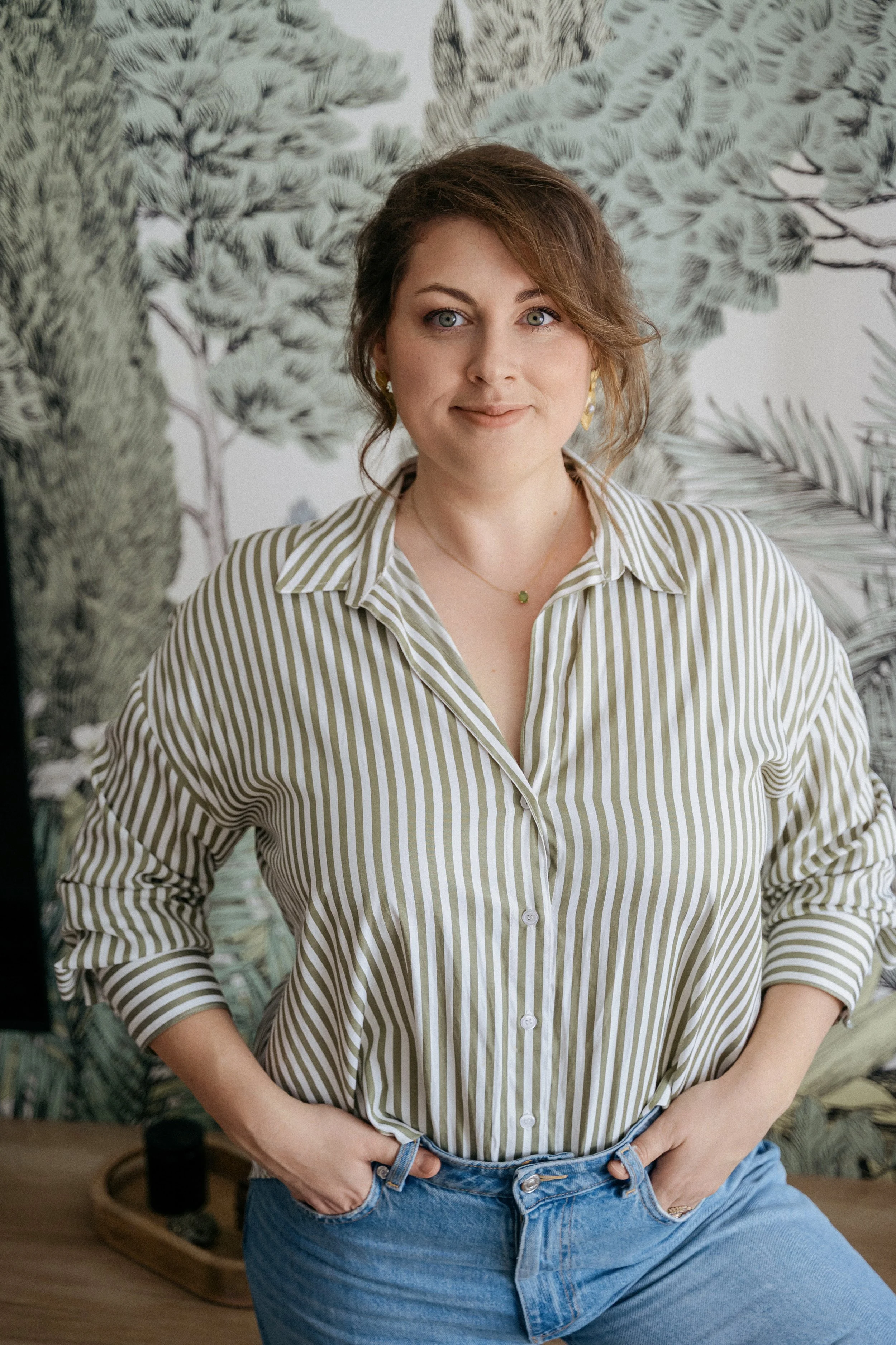 Une jeune femme avec des cheveux bruns ondulés et des yeux bleus, portant une chemise à rayures beige et blanche, et un jean bleu, posant devant un mur de décoration avec des dessins d'arbres et de feuillage.