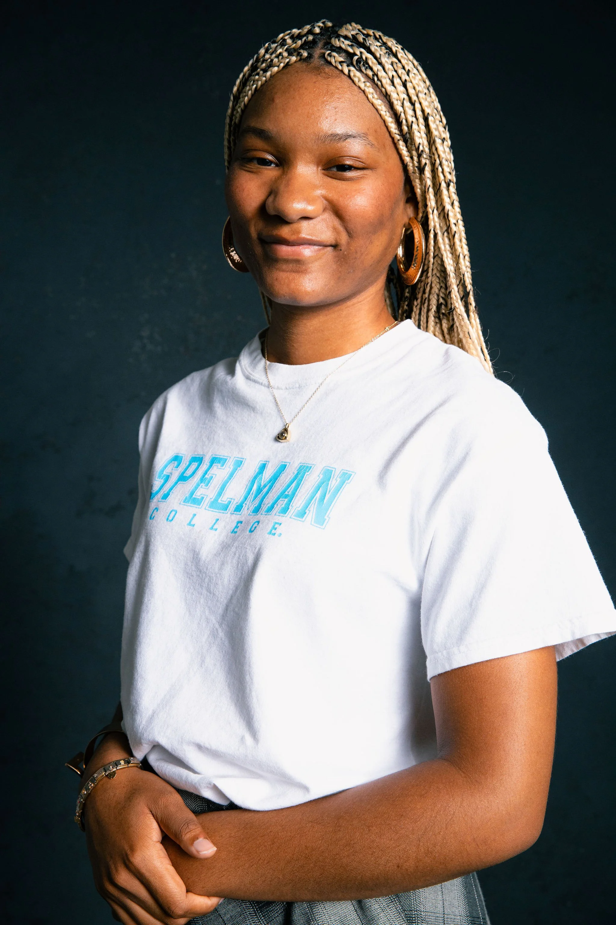 A young woman with braided blonde hair wearing a white T-shirt with blue text, gold hoop earrings, a necklace, and a watch, standing against a dark background.