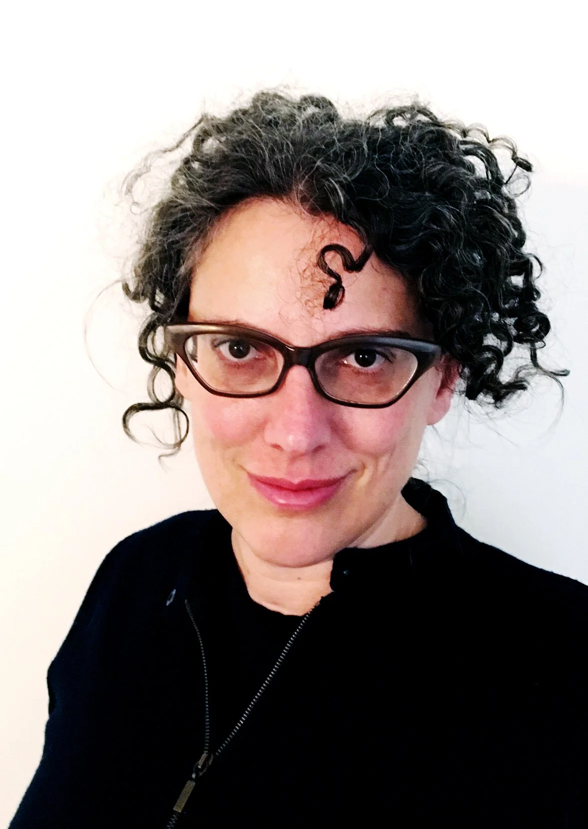 Woman with short curly dark hair, wearing glasses and a black jacket posing against a plain white background.