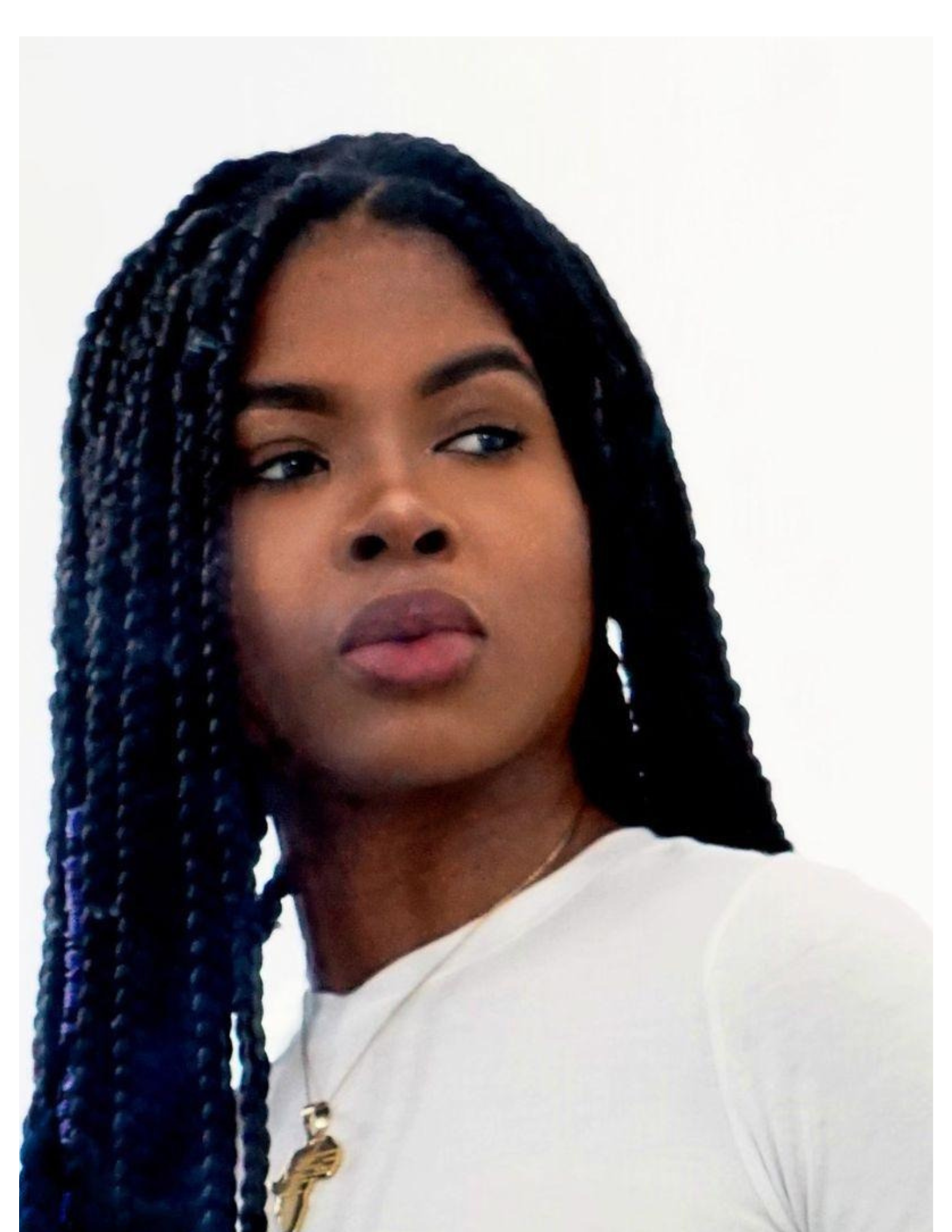 Close-up of a young woman with long braided hair, wearing a white top, gold necklace, and neutral makeup, looking to her left against a plain background.