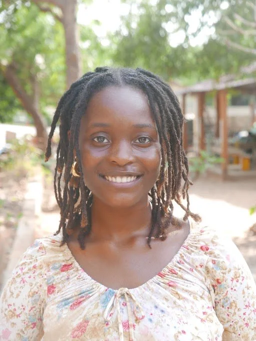 A smiling woman with shoulder-length dreadlocks, wearing a floral blouse and gold hoop earrings, outdoors in a green, wooded area.