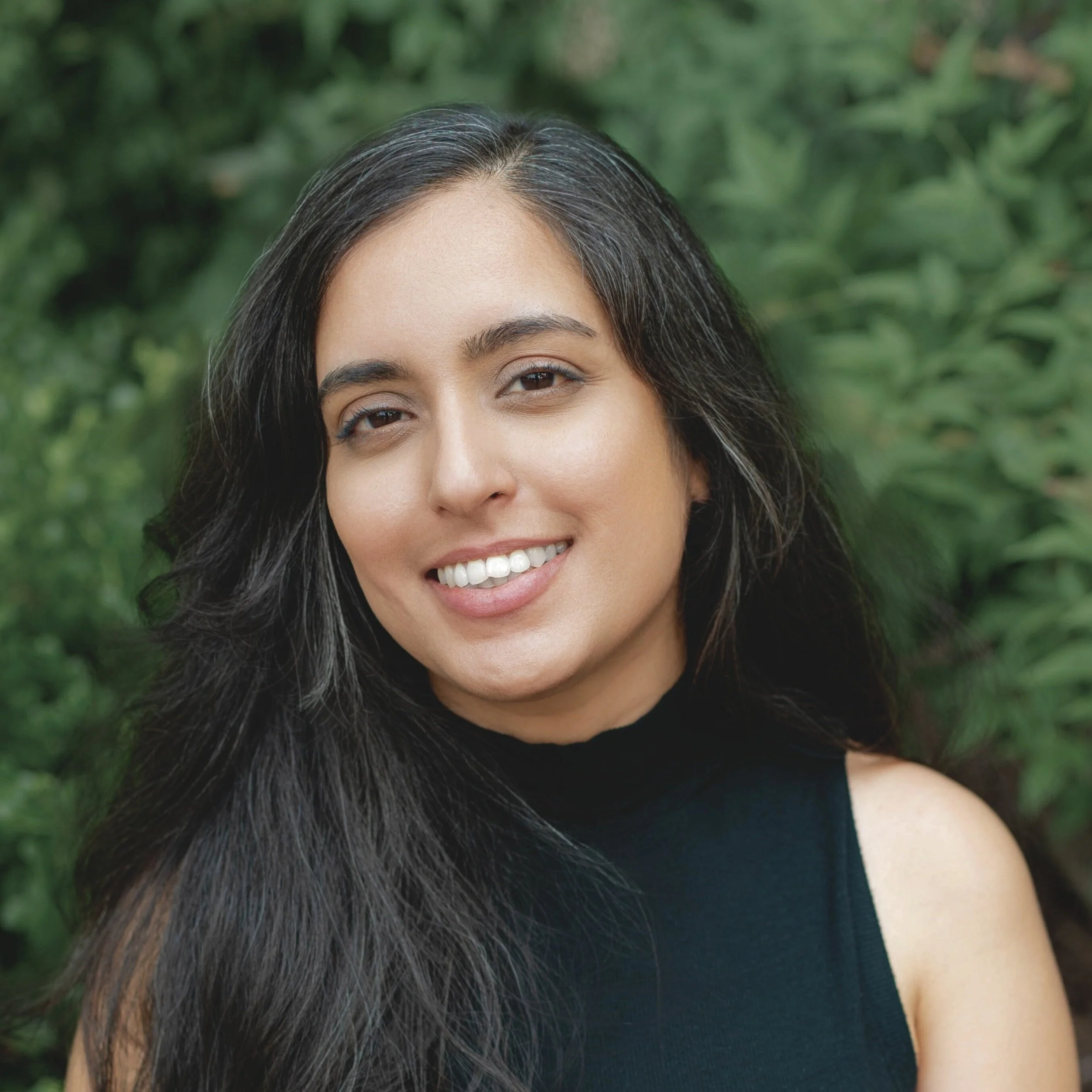 A woman with long dark hair smiling outdoors against a background of green foliage.