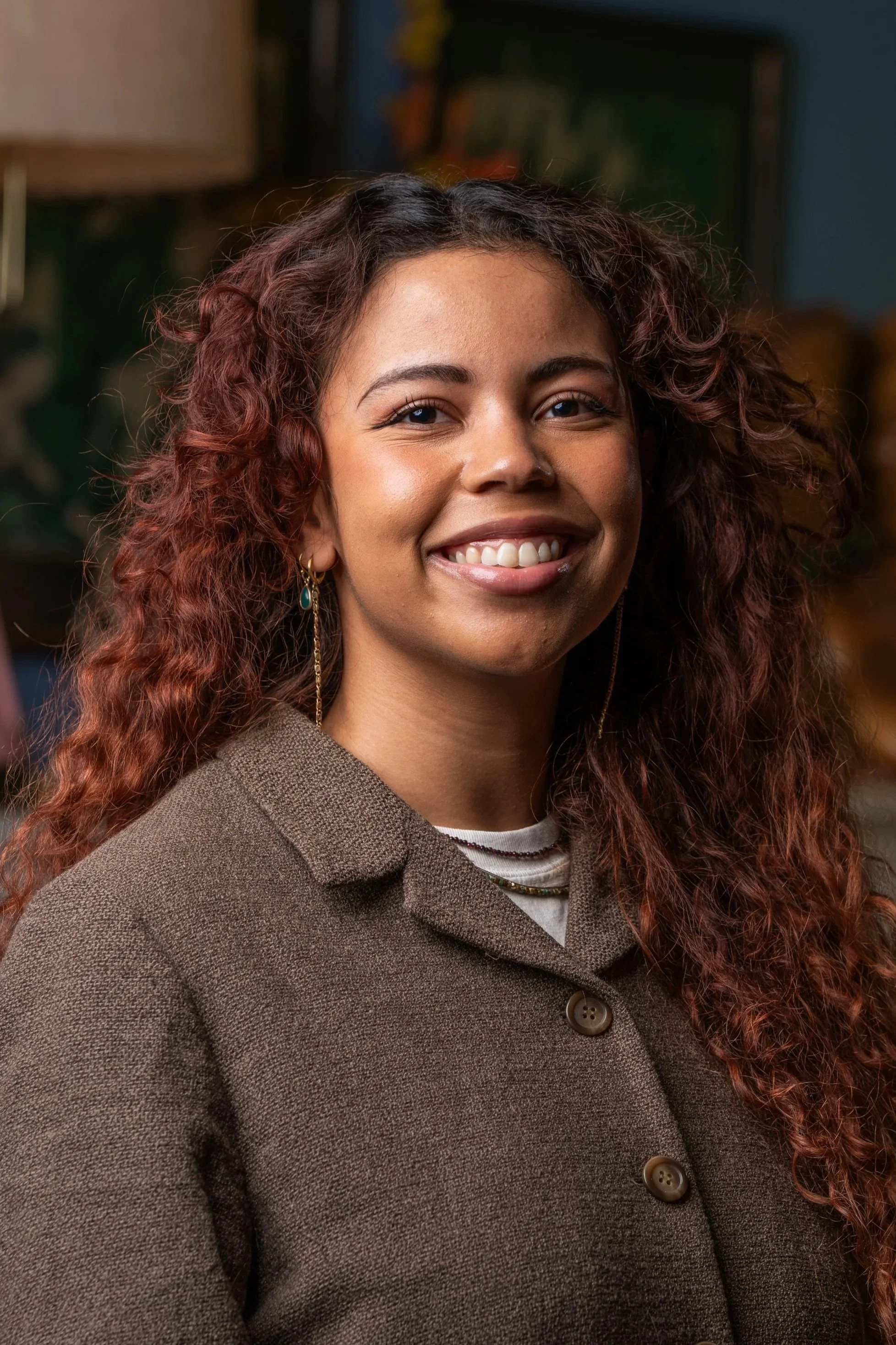 A woman with long, curly, ombré hair smiling outdoors in front of blooming tree branches.