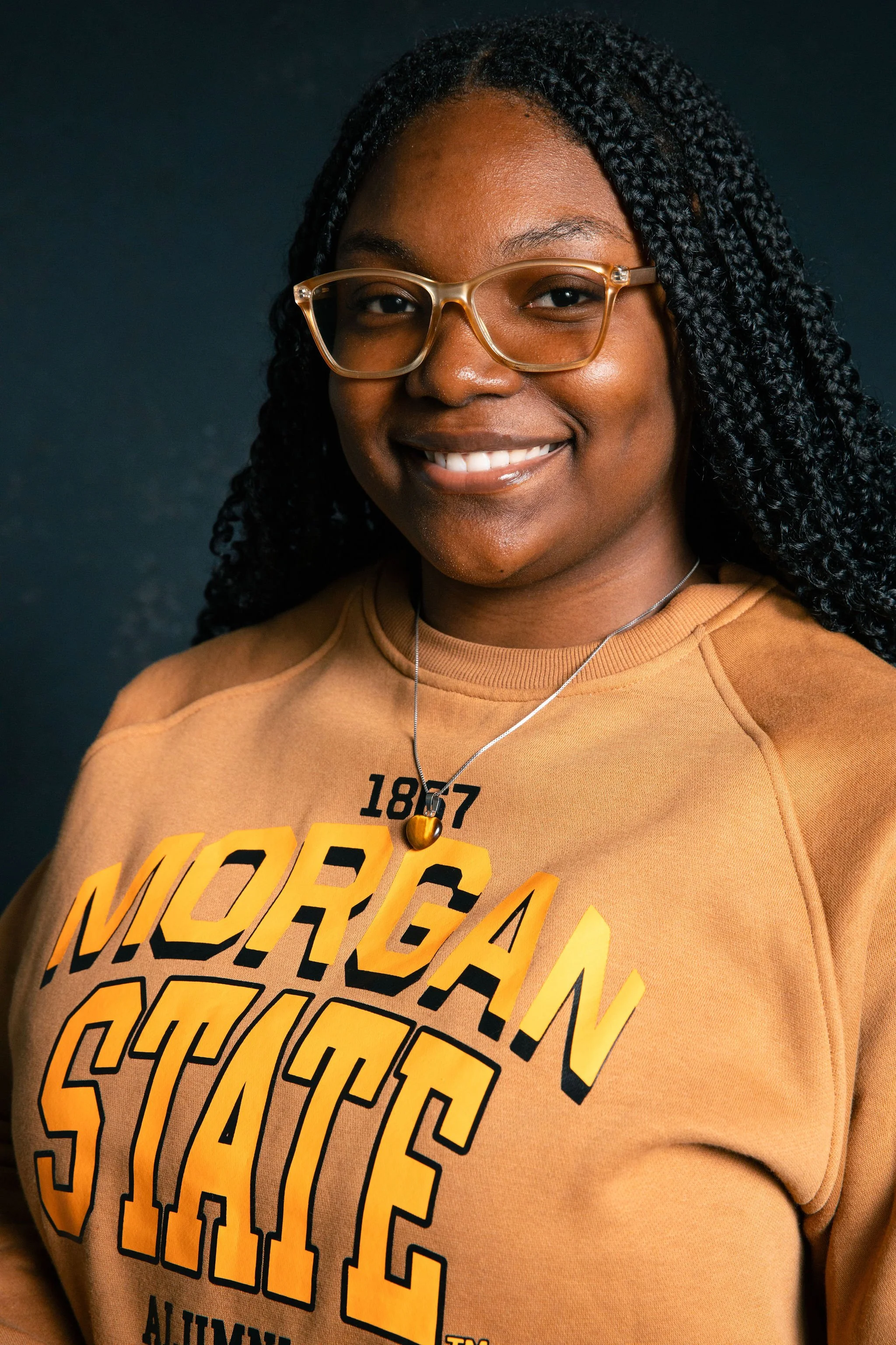 A woman with dark curly hair and glasses, smiling, wearing a Cornell University sweatshirt with the text "Morgan State" on it.