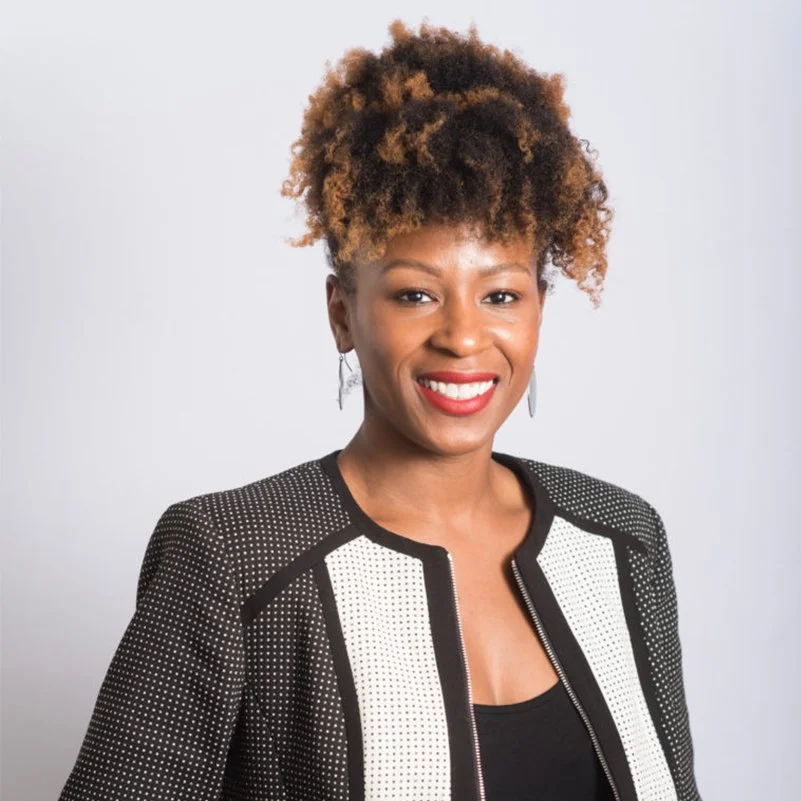 A smiling woman with short, curly, ombré hair wearing a black and white patterned jacket and earrings, standing against a plain white background.