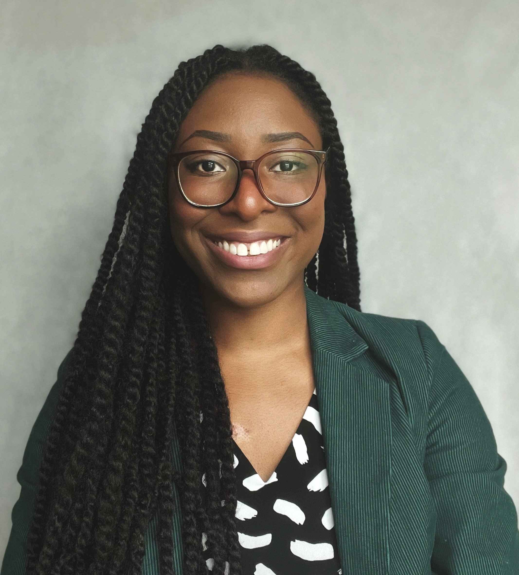 A young woman with long braided hair, wearing glasses, a green blazer, and a patterned top, smiling at the camera against a light grey background.