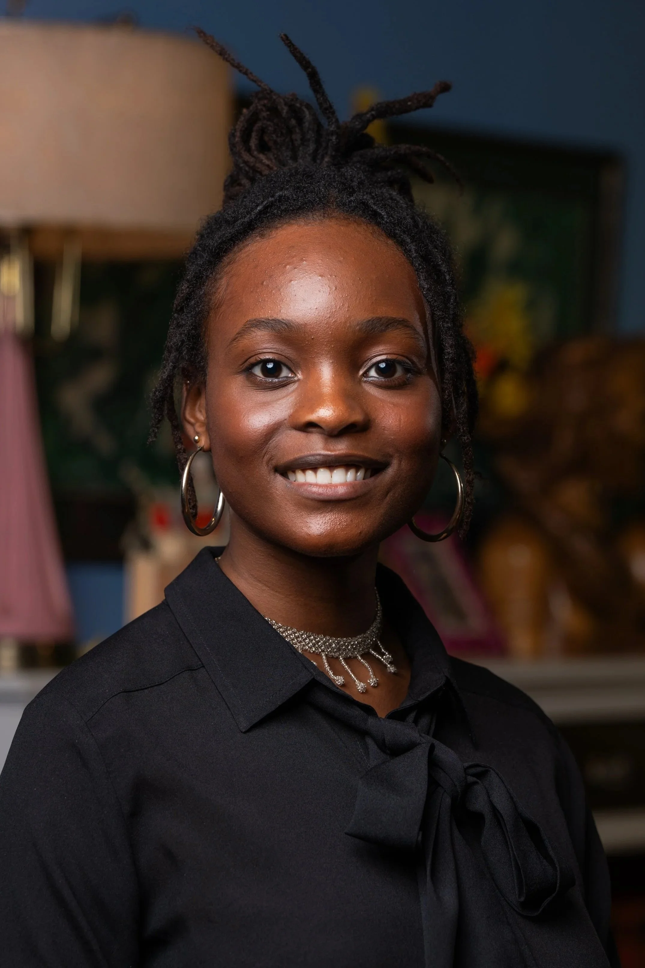 A smiling woman with shoulder-length dreadlocks, wearing a floral blouse and gold hoop earrings, outdoors in a green, wooded area.