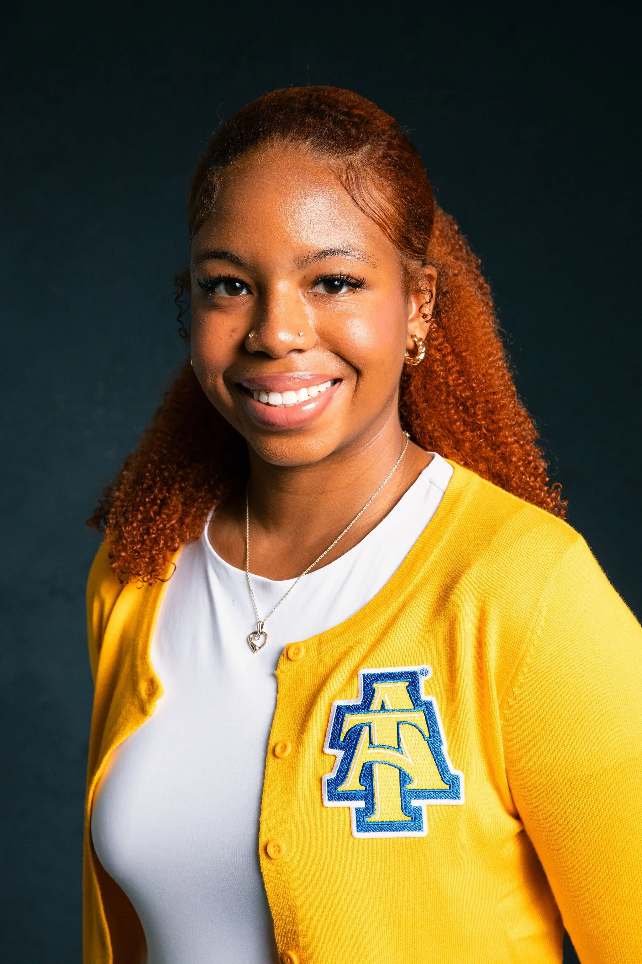A young woman with curly red hair, smiling, wearing a yellow cardigan with a blue and yellow embroidered logo, against a dark background.