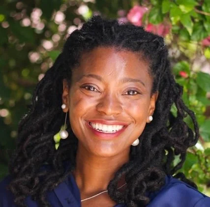 A smiling woman with dark braided hair and pearl earrings outdoors with greenery and pink flowers.