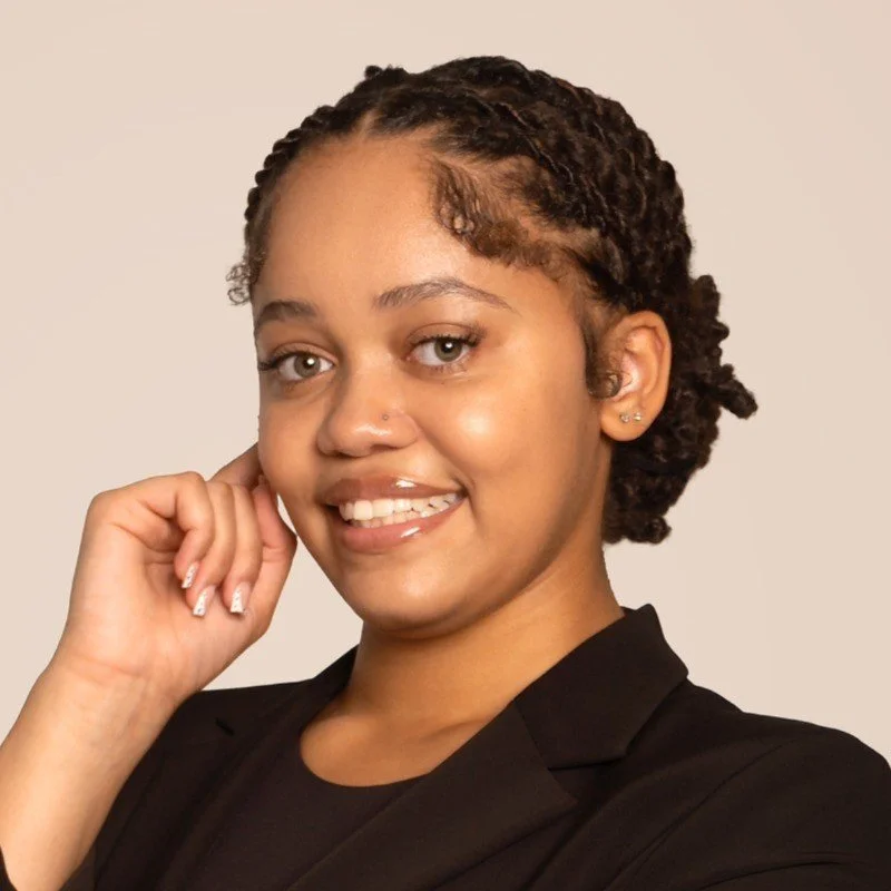 A woman with short, curly hair is smiling and posing with her hand near her face, wearing a black blazer against a light background.