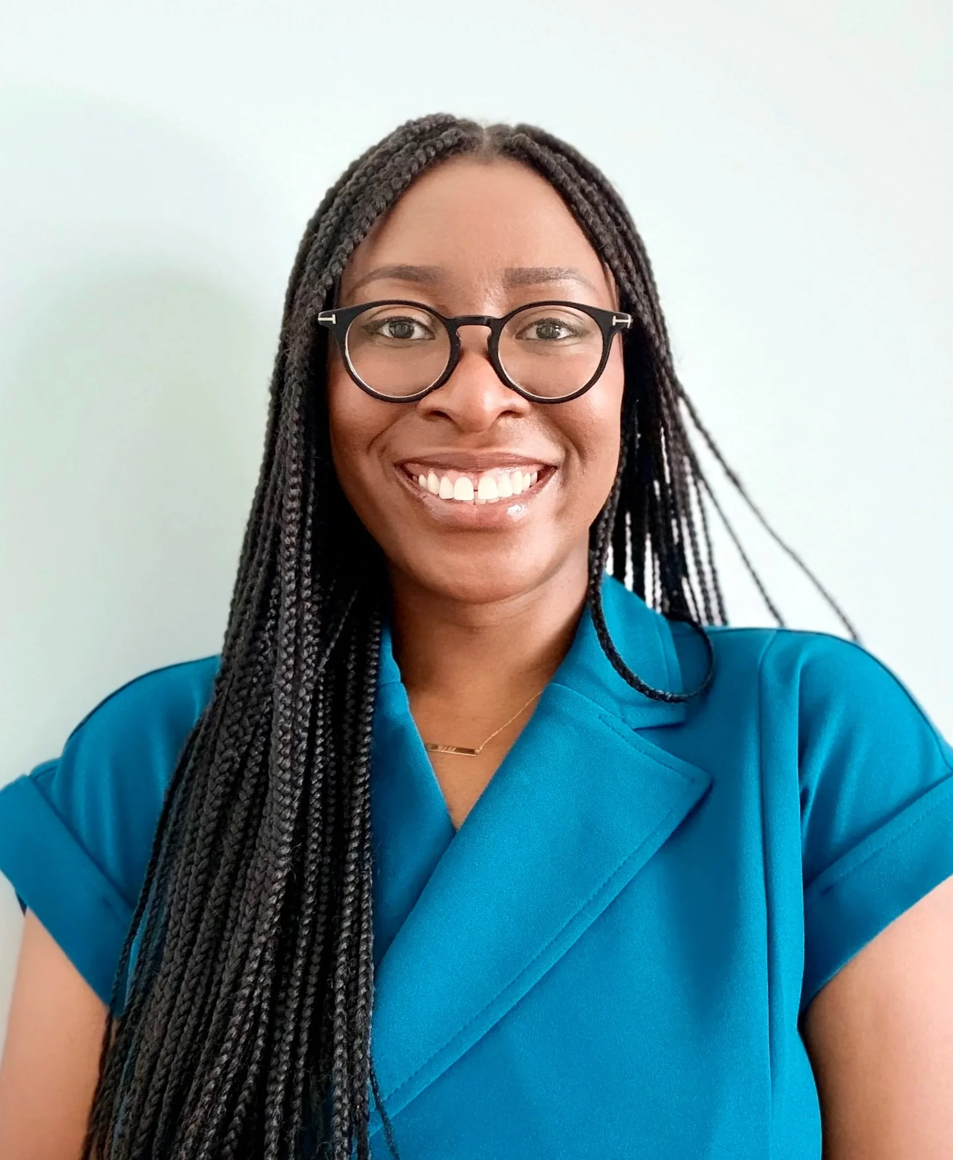 Portrait of a smiling woman with glasses, long braided hair, wearing a dark green blazer over a black and white patterned top, standing against a plain light-colored background.