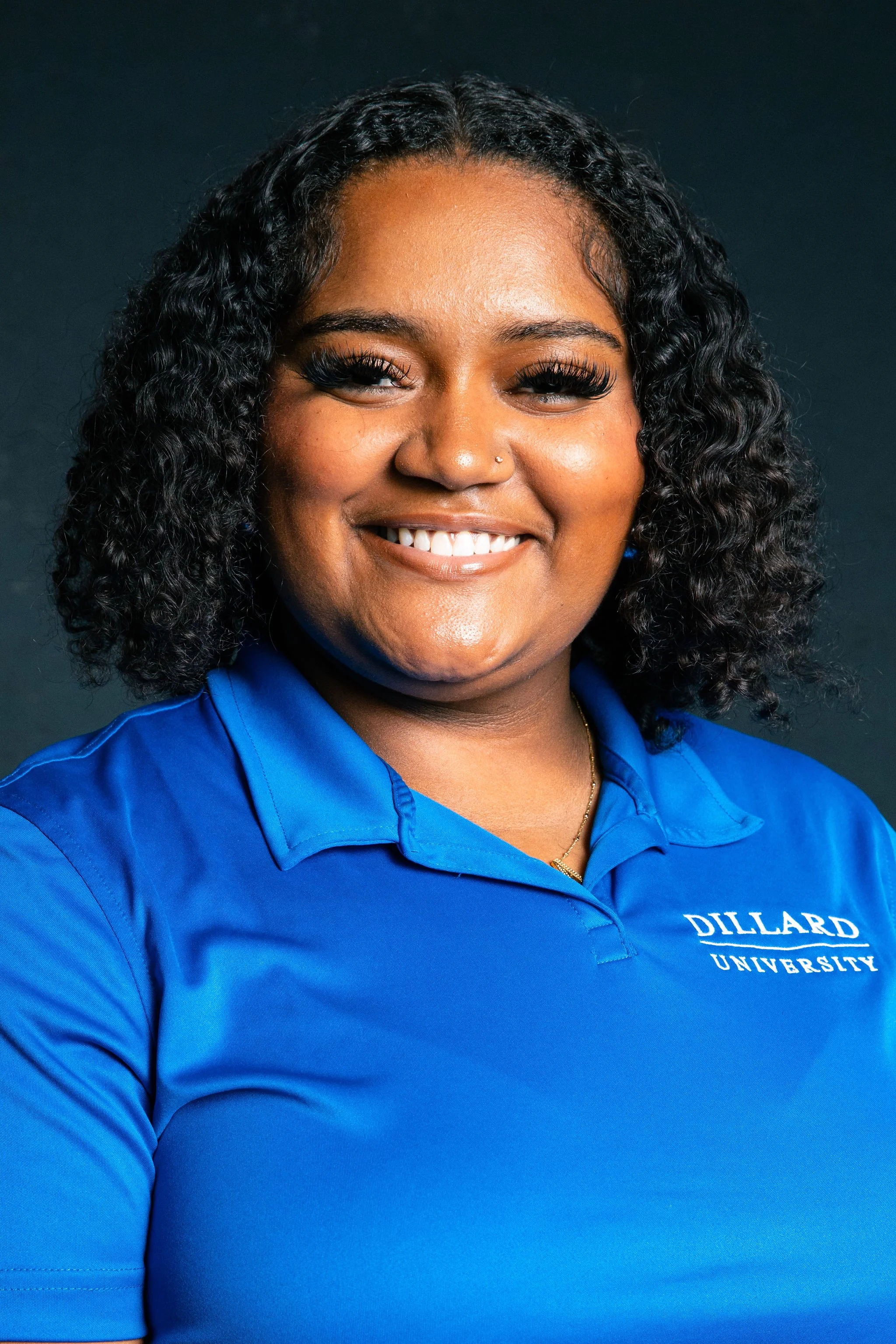 Smiling woman with curly black hair wearing a blue polo shirt with 'Dillard University' embroidered on it, against a dark background.