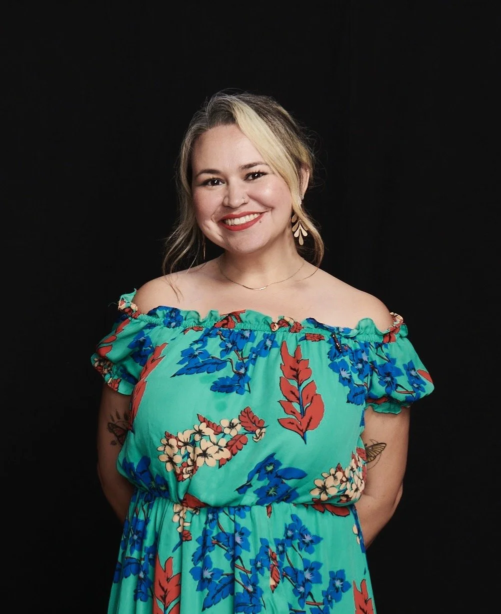 A woman with blonde and brown hair, wearing a vibrant turquoise floral off-the-shoulder dress, smiling at the camera, standing against a black background.