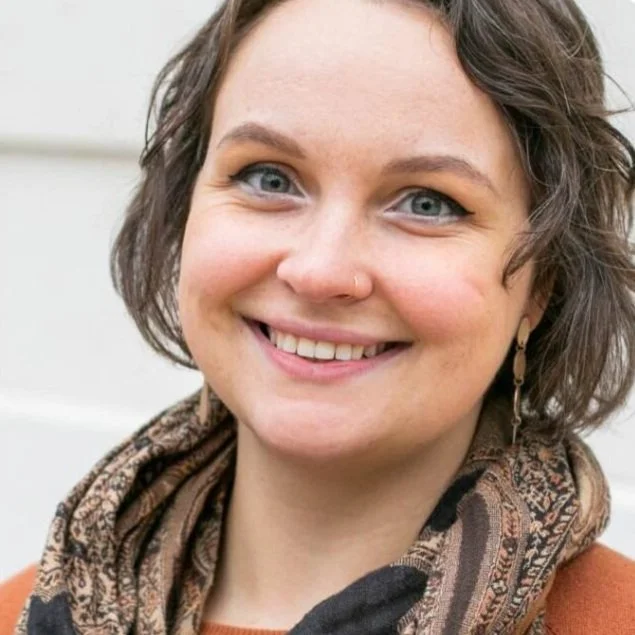 Close-up of a smiling woman with short wavy brown hair, blue eyes, a nose piercing, earrings, and a patterned scarf.