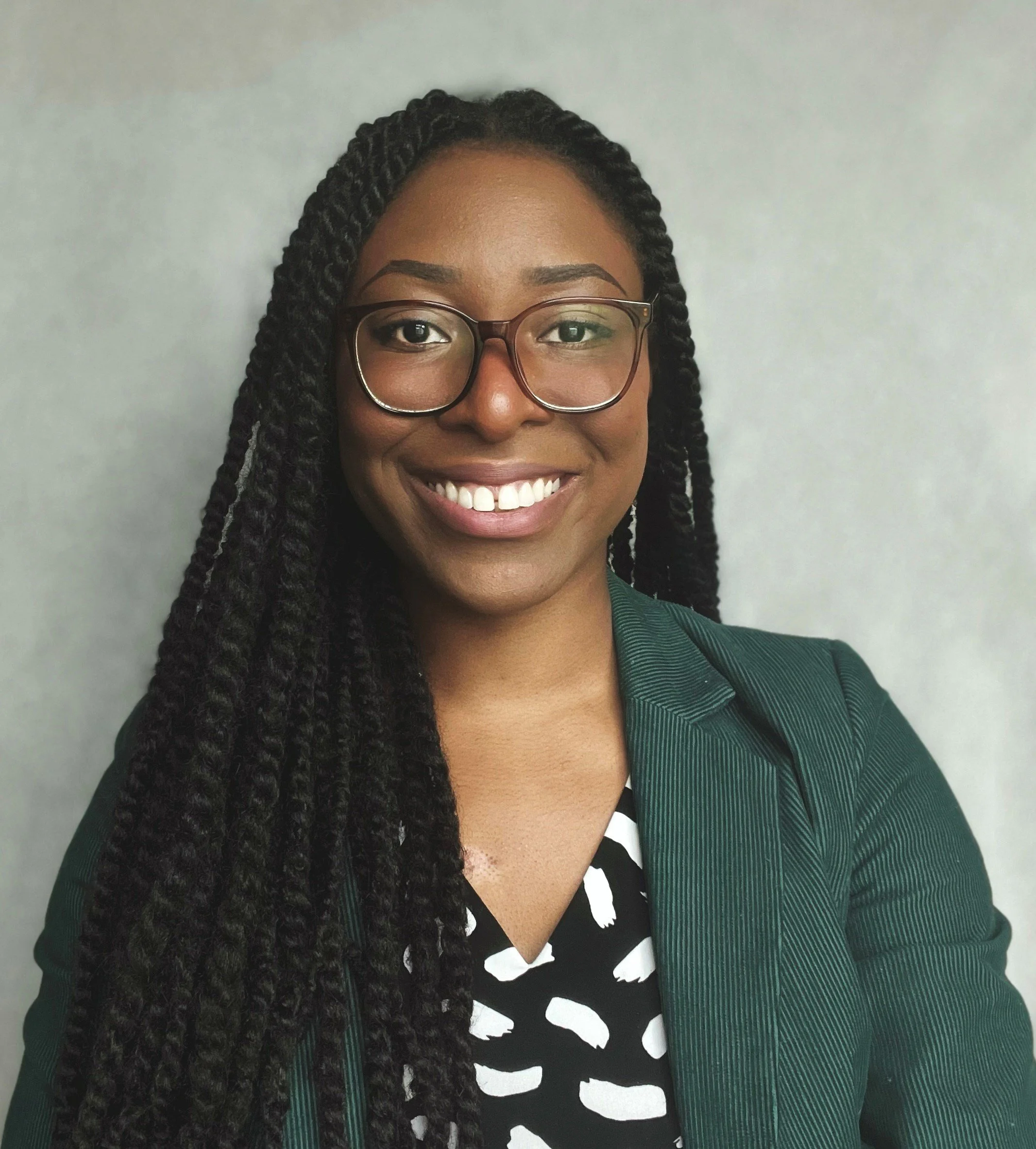Portrait of a smiling woman with glasses, long braided hair, wearing a dark green blazer over a black and white patterned top, standing against a plain light-colored background.