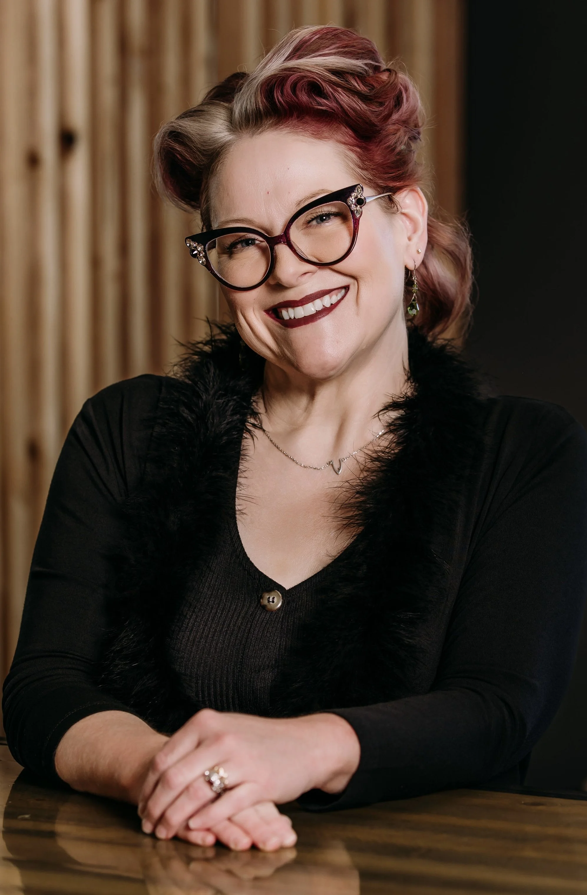 A woman with pink and brown hair, wearing glasses with decorative rhinestones, smiling, sitting at a table with crossed hands, against a wooden background.