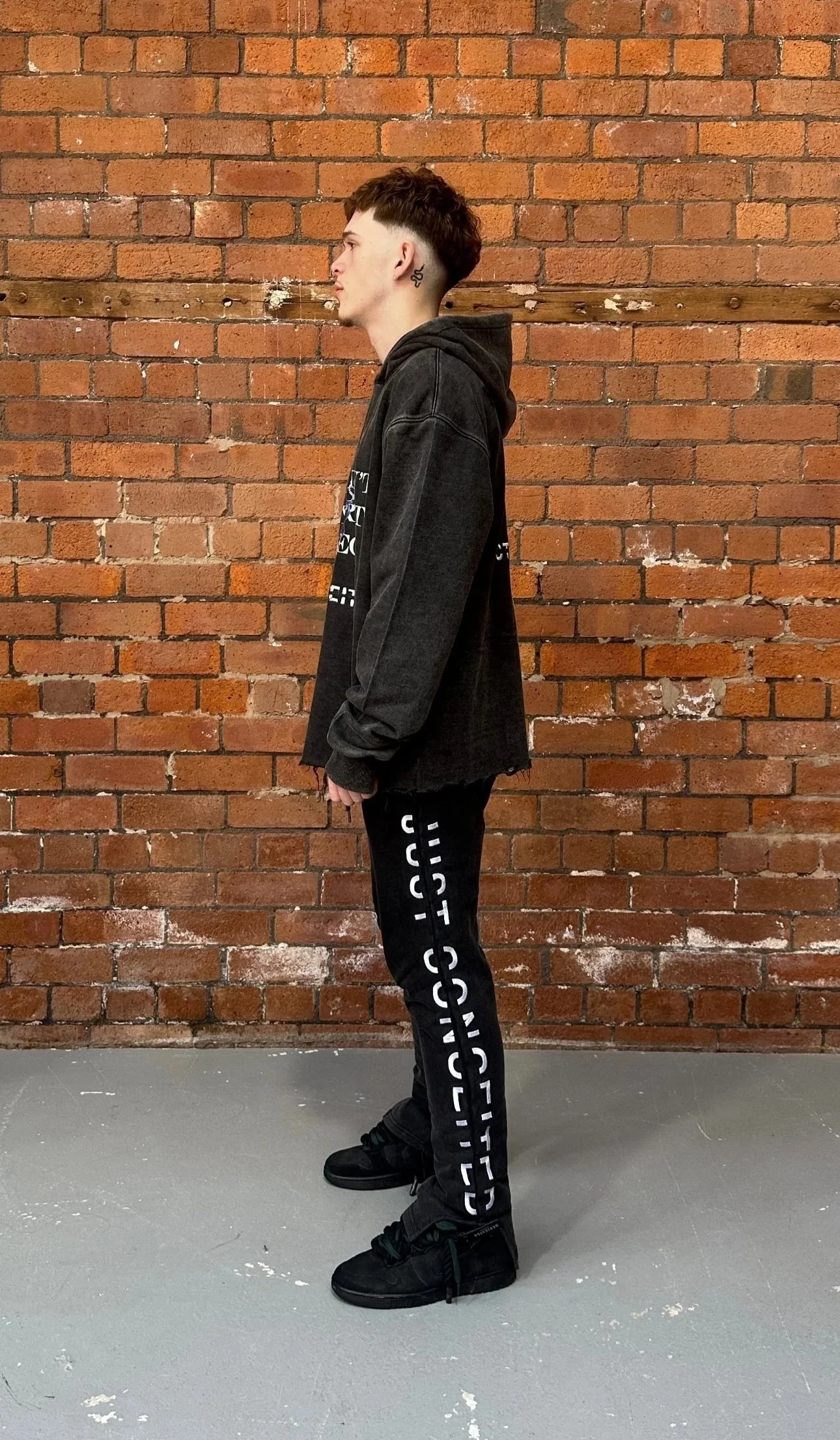 Side view of a young man with short, dark, and slightly curly hair standing against a brick wall. He is wearing a dark hoodie, black track pants with bold white text on the side, and black sneakers.