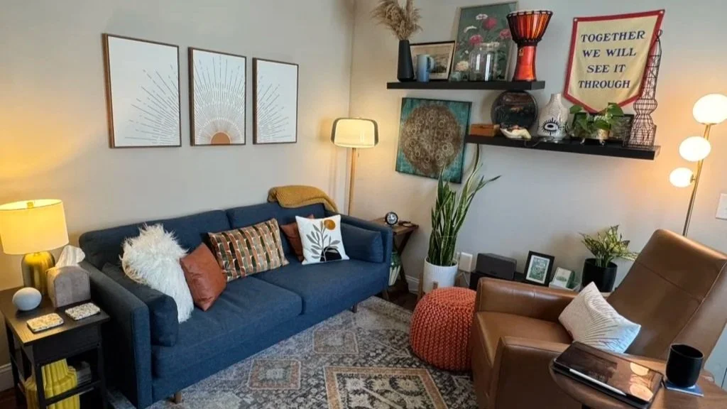 A warm therapy office with a navy sofa, leather armchair, layered shelving, plants, and a banner reading Together We Will See It Through