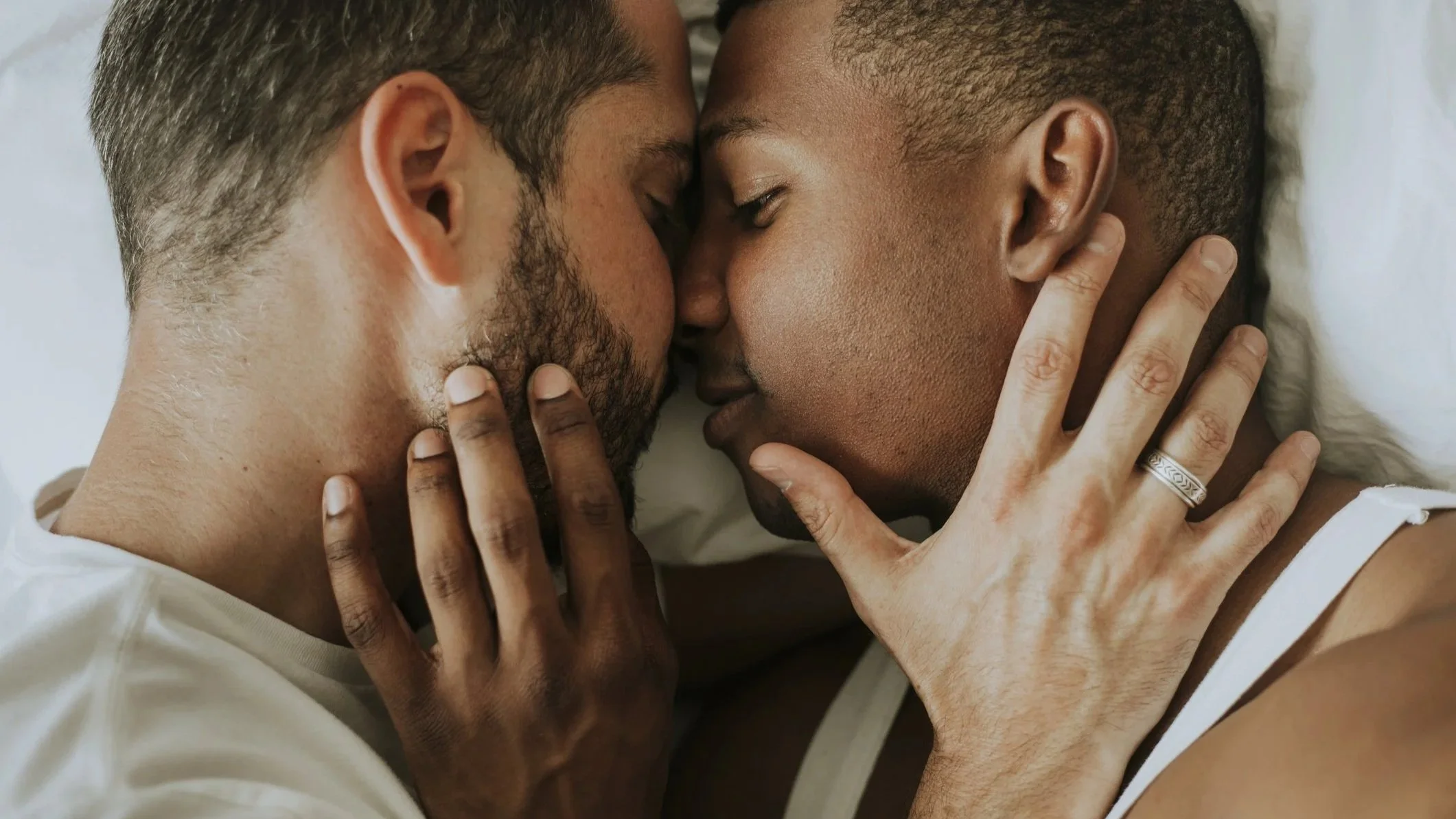 Two men cuddle in bed, lying on their sides nose to nose, each placing a hand on the other’s neck and chin.