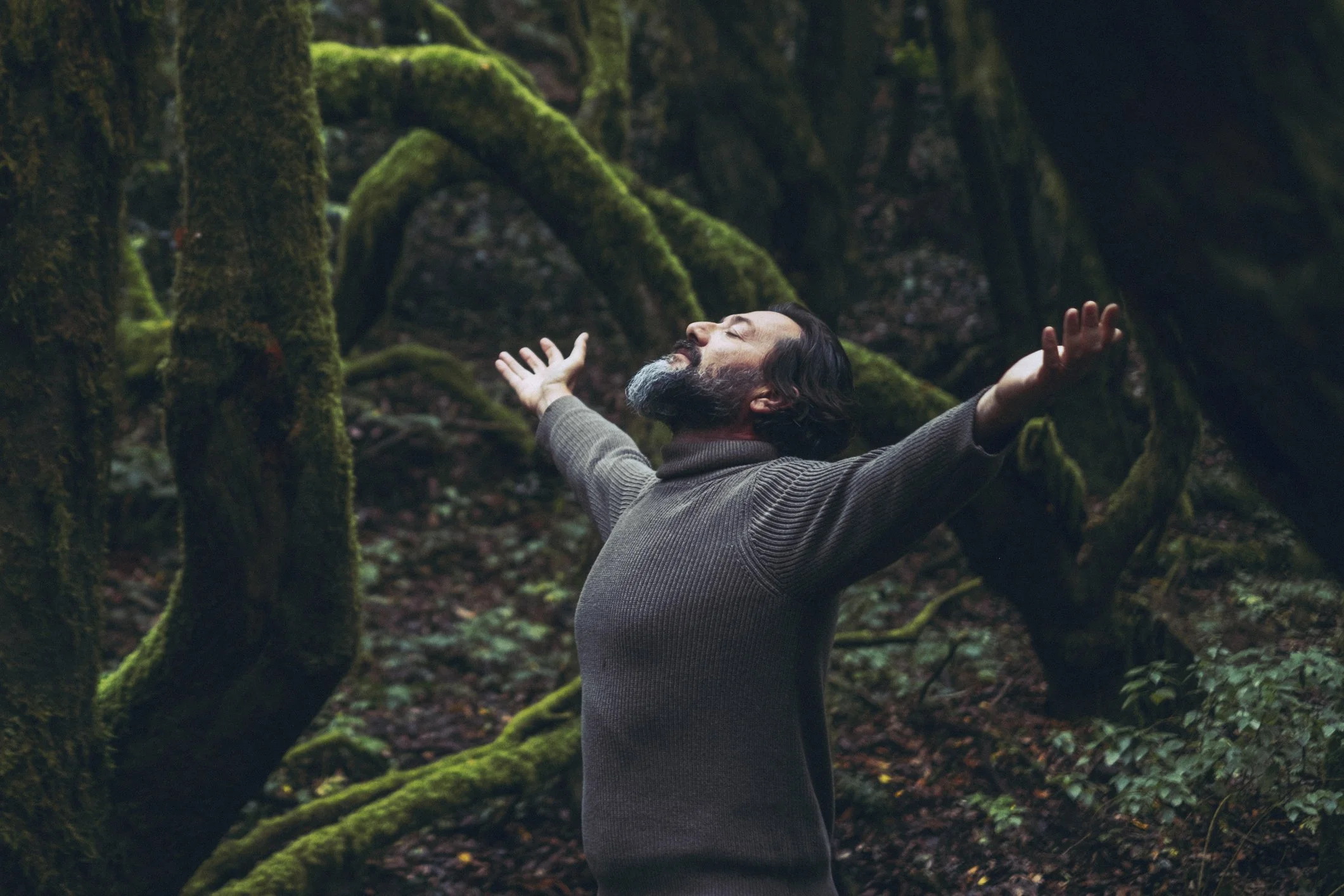 A man with a graying beard stands in a moss-covered forest, eyes closed, head tilted back, arms outstretched, surrounded by moss-draped tree trunks and roots in dim natural light.