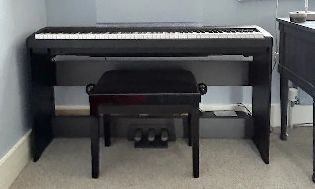 A black digital piano on a stand with a matching black bench, positioned against a light blue wall in a room with gray carpet.
