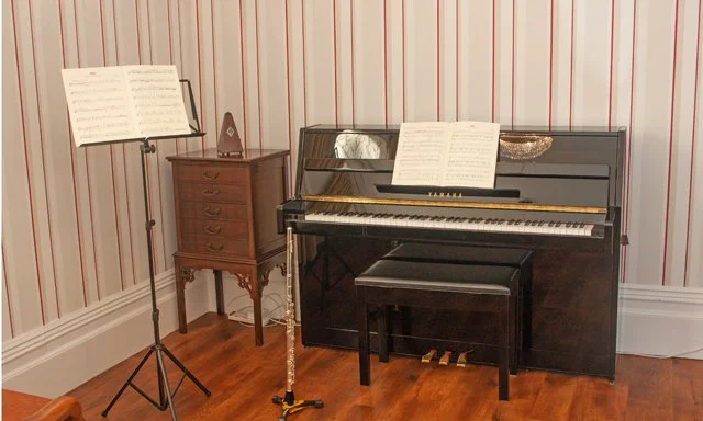 A black upright piano with a matching bench, with sheet music on the music stand, situated in a room with striped wallpaper and wooden flooring. To the left, there is a small wooden table with open sheet music and a triangular metronome.