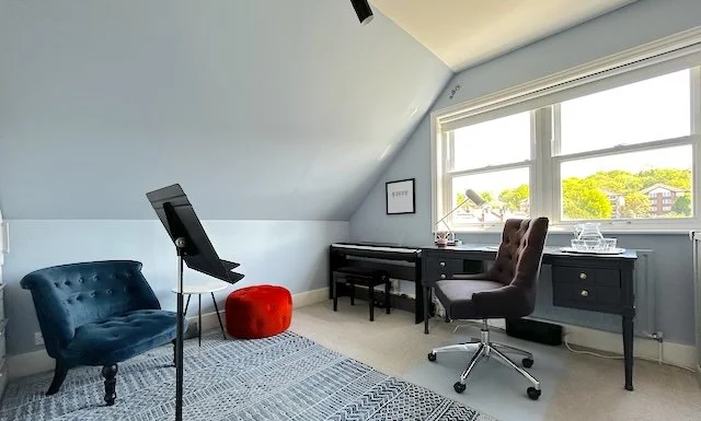 Home office with a large window, gray walls, a black desk, a gray swivel chair, a blue tufted armchair, a red ottoman, and a music stand.