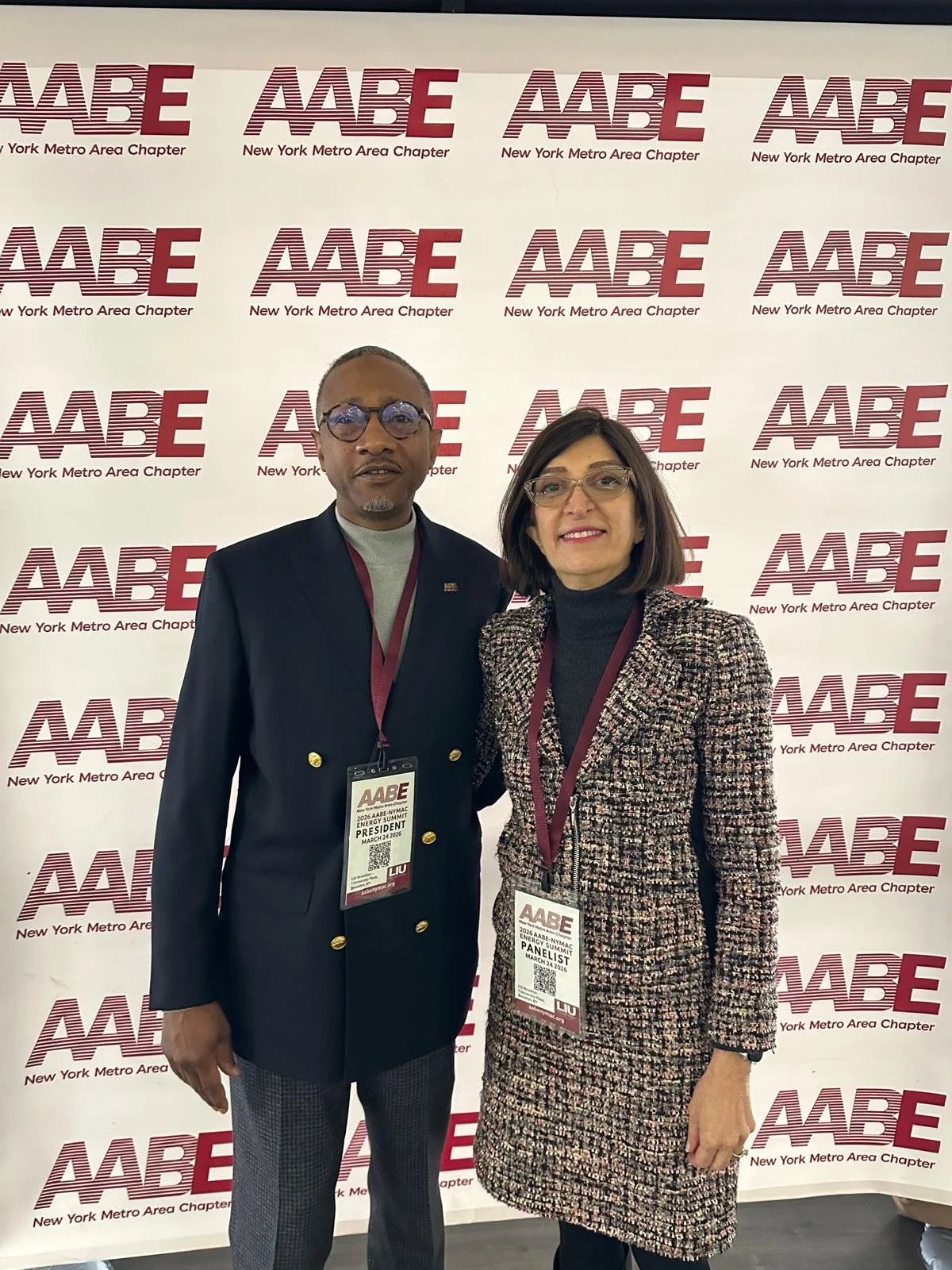 Two individuals standing in front of a backdrop with the AABE logo repeatedly printed, at an event. The man on the left is wearing a dark blazer, a light gray shirt, and checkered trousers. The woman on the right is dressed in a tweed jacket and black top, both wearing event badges around their necks.