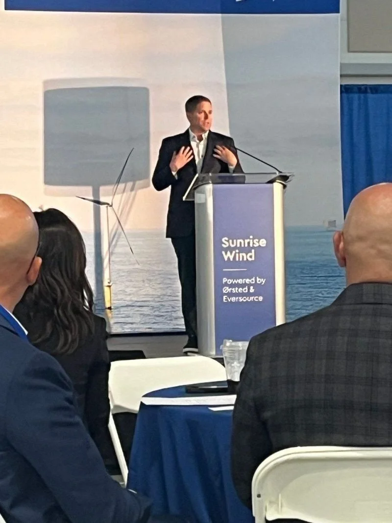 A man in a dark suit is speaking at a conference podium labeled 'Sunrise Wind, Powered by Ørsted & Eversource.' He appears to be gesturing with his hands while addressing an audience in a conference room.