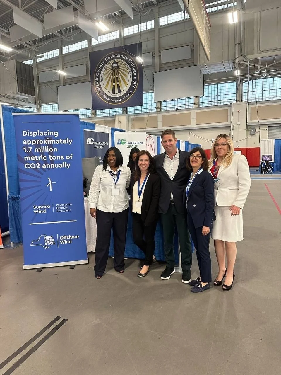 Group of five people standing together at a conference in an indoor exhibition hall, smiling at the camera, with a blue display sign about CO2 displacement and wind power projects in the background.