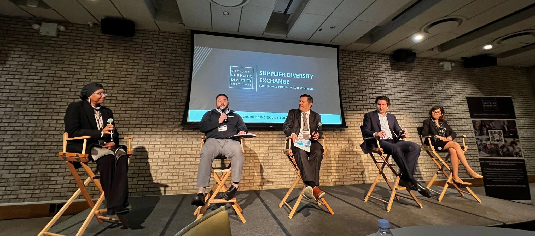 A panel of five people sitting on risers during a discussion event about supplier diversity exchange. Four of the panelists are holding microphones, and there is a large screen behind them displaying the event details.