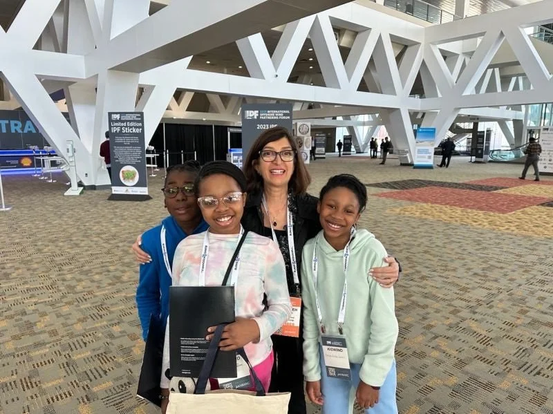 Group of four people, three children and one adult woman, smiling and posing for a photo at a conference or convention center.