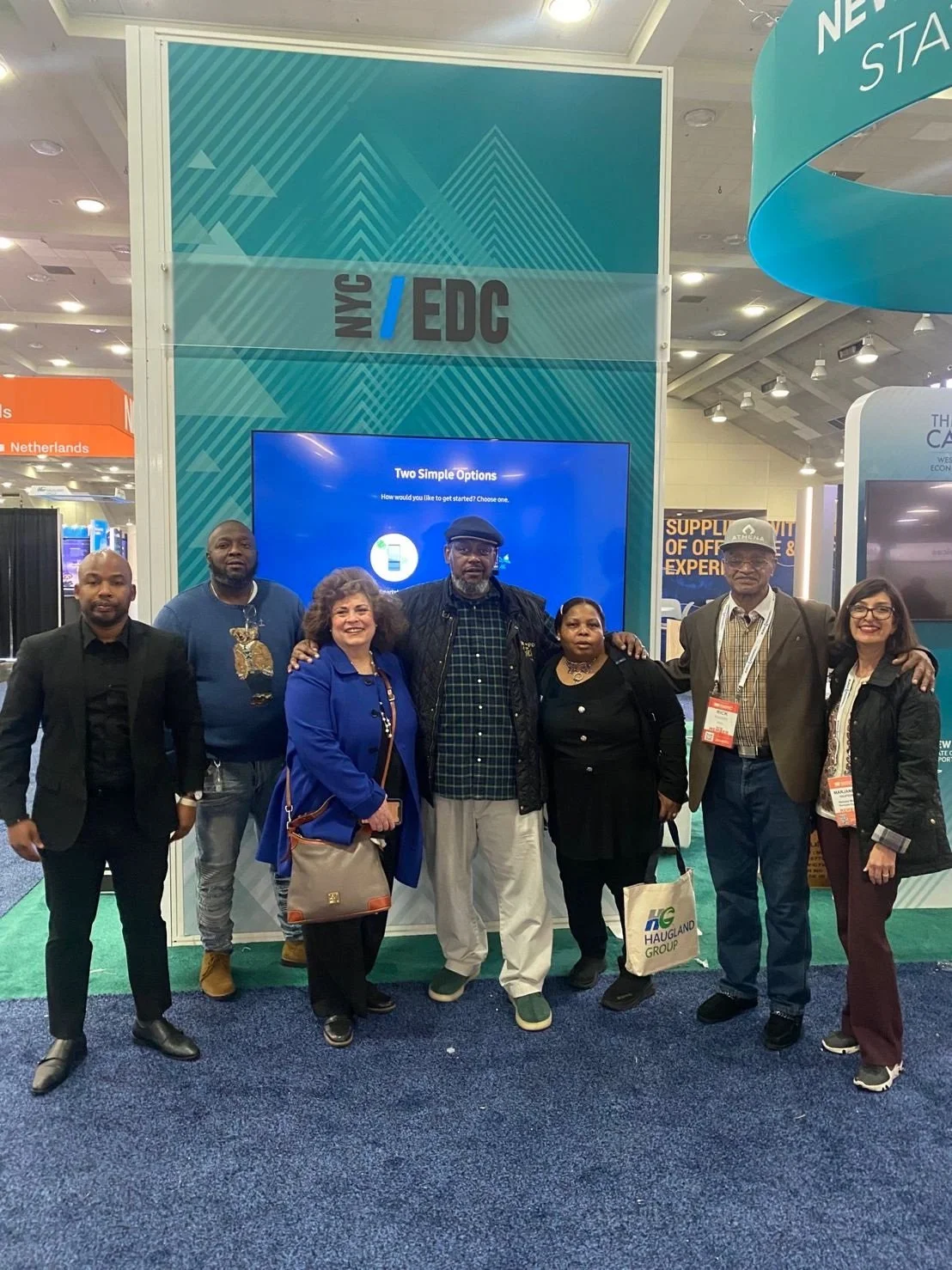 Group of seven people standing together at a conference booth with a blue and teal backdrop, displaying the text 'NYC / EDC' and a screen showing options for starting a process, with conference badges around their necks.