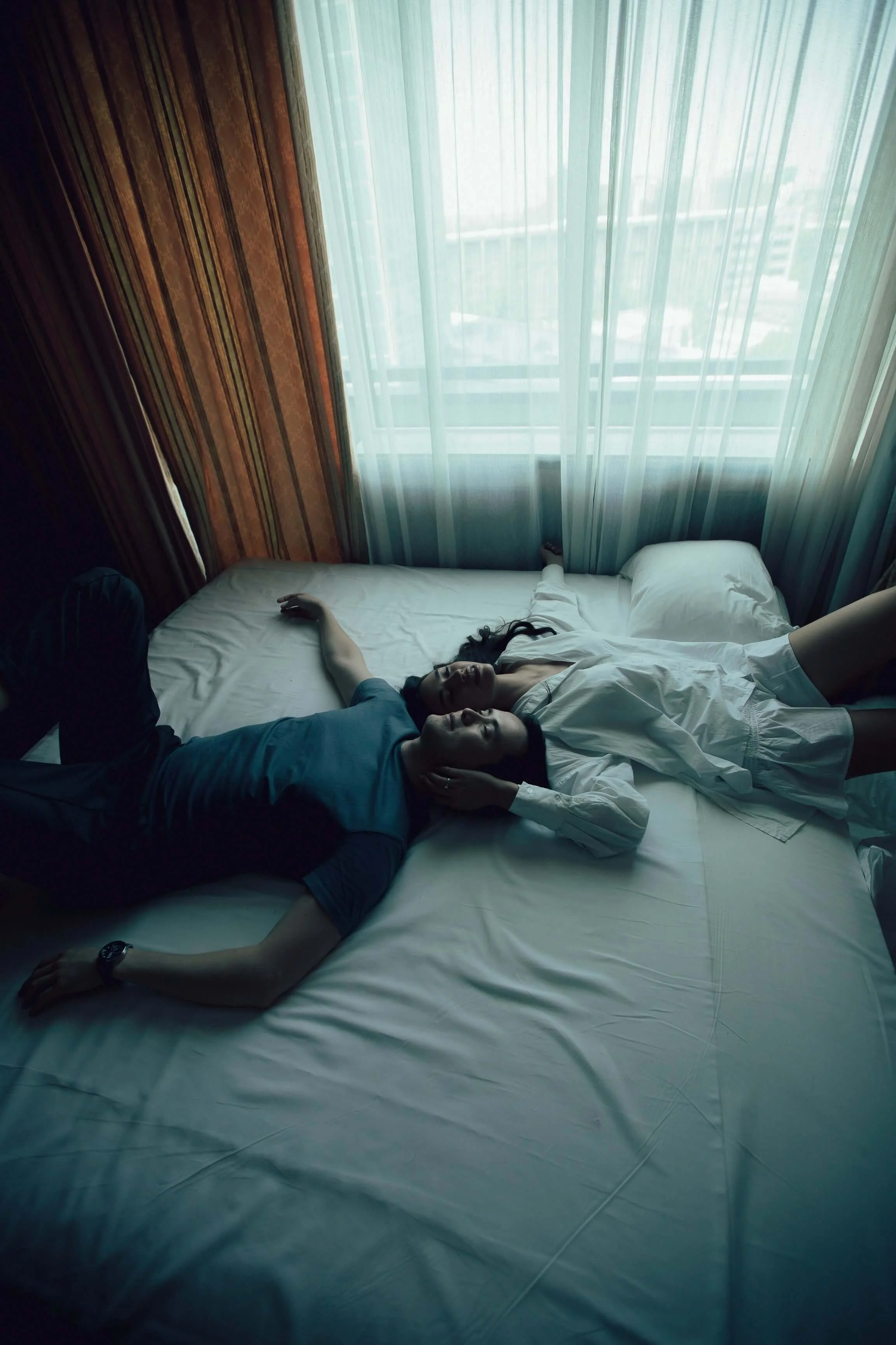 Two people lying on a bed after a therapy session near a large window with sheer curtains, one with arm extended, both appearing relaxed.