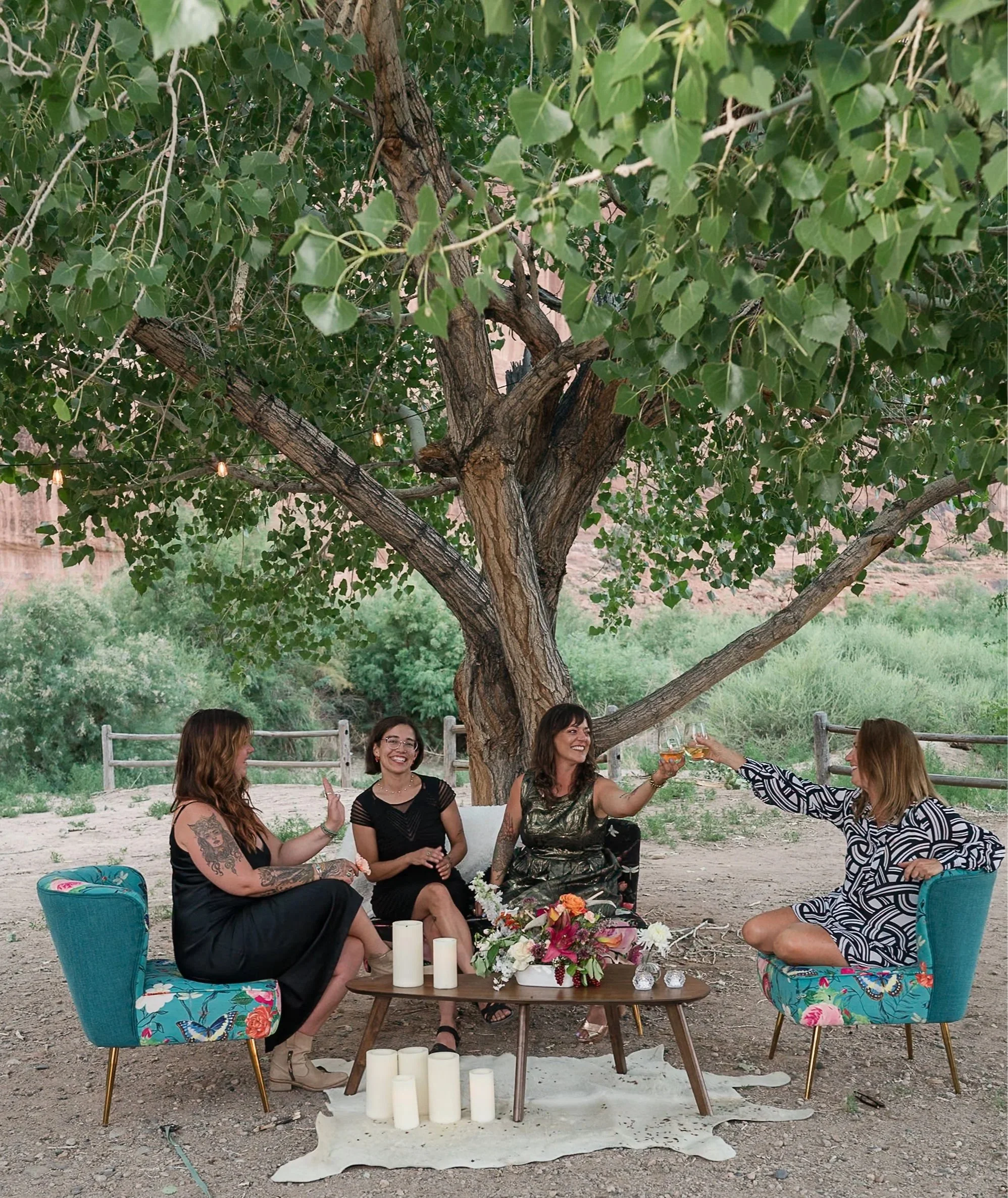 Four women sit under a tree in the desert of Moab Utah enjoying cocktails at a corporate event.