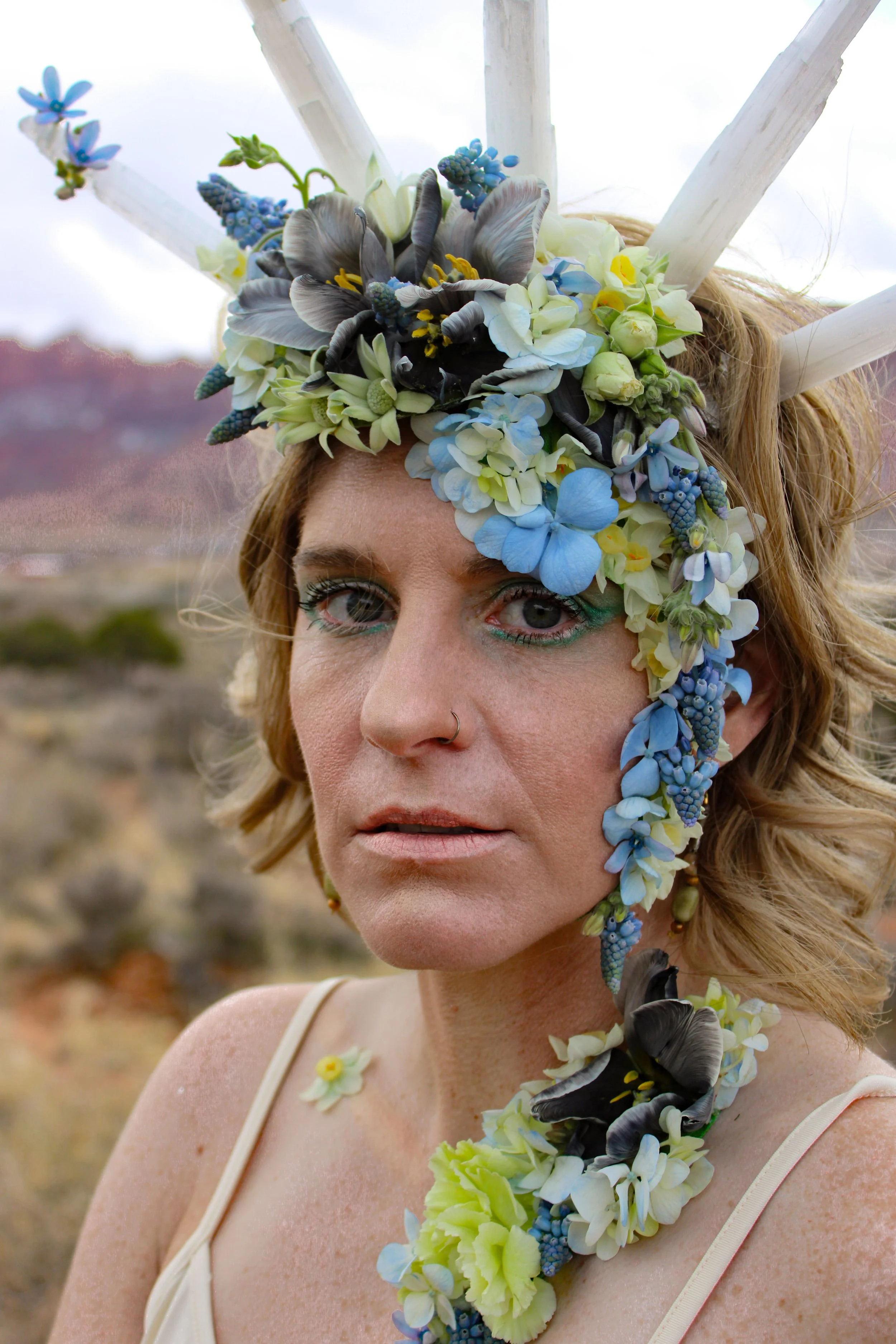 Woman wearing a selenite crown with flowers cascading down her face and neck looks directly into the camera as she stands on a desert landscape with wearable floral art.