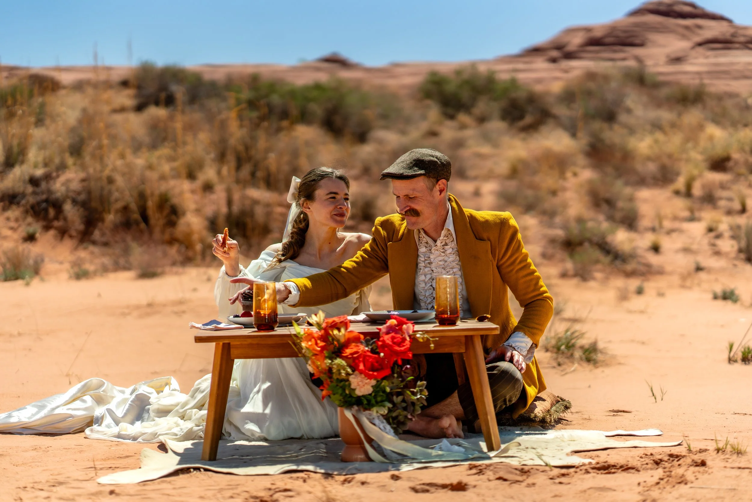 A bride and groom sit on a low picnic table with plates and glasses and a lovely bouquet of flowers in front of them in the wild Moab desert