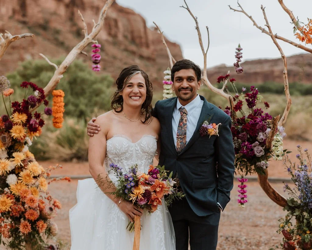 Bride and groom stand in front of colorful florals in the style of an Indian wedding in the Moab Desert at the Red Earth Venue