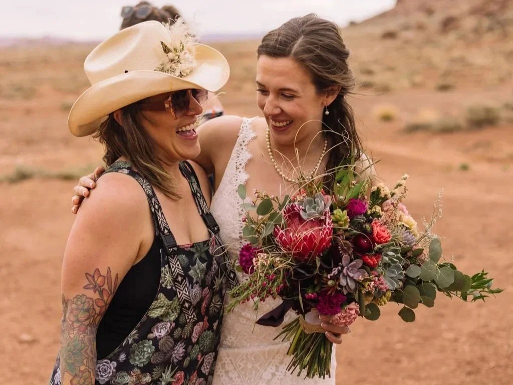 Toni from Tangled Sage hugs a bride holding a big pink and purple bouquet with succulents and protea outside at The Red Earth Venue in Moab Utah