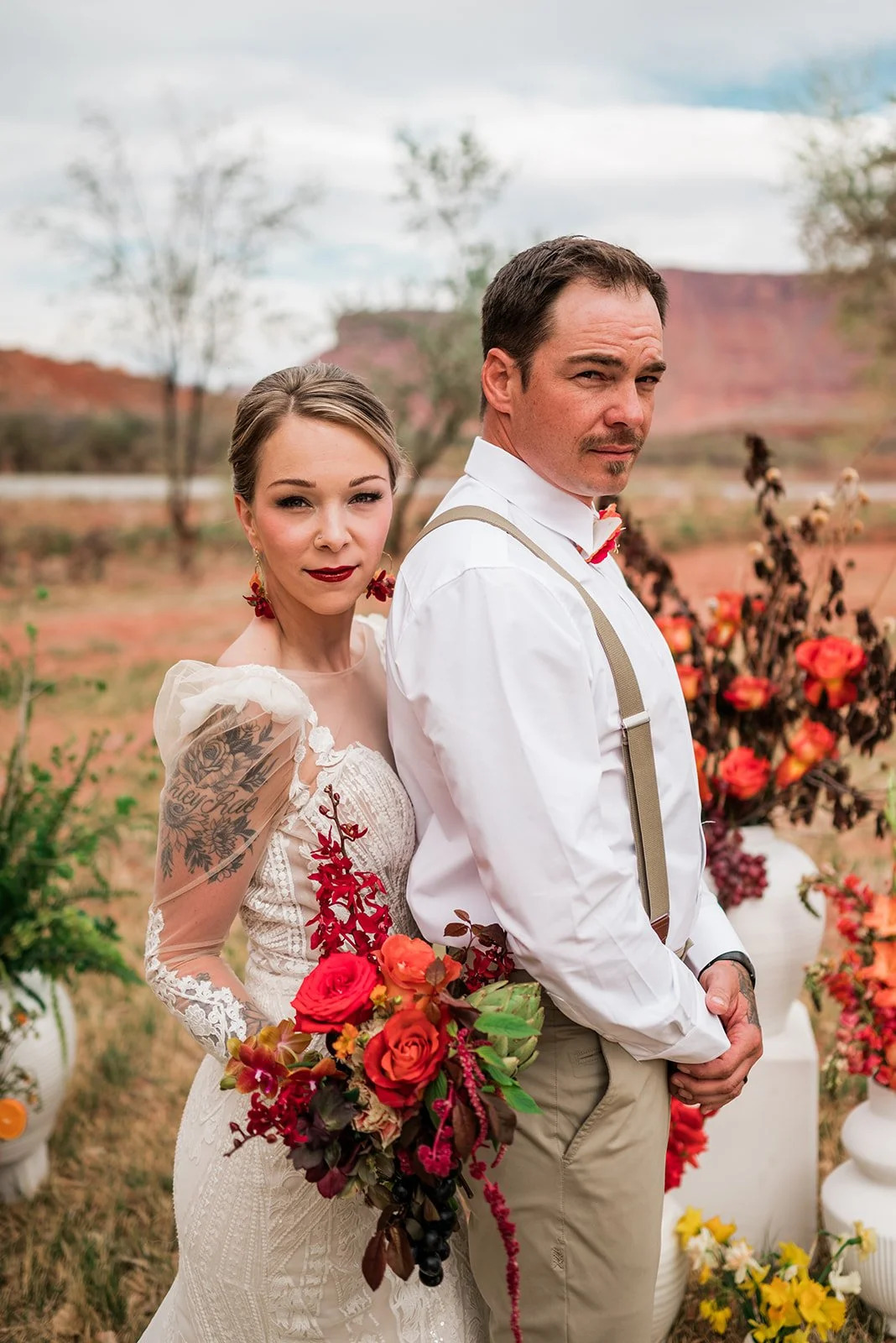 Bride and groom pose in front of the Colorado River at Red Cliffs Lodge in Moab Utah holding brightly colored flowers.