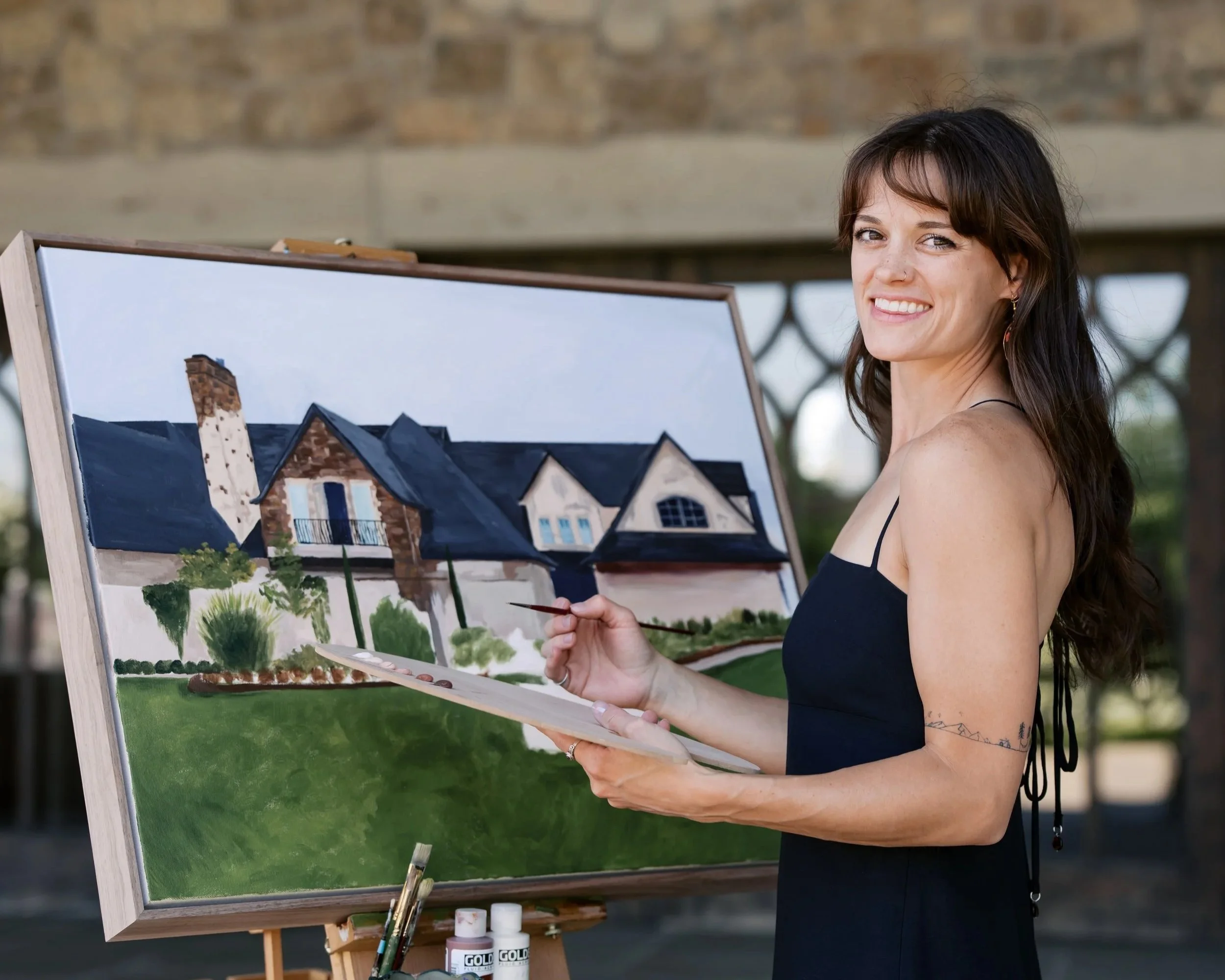 A woman with dark hair, a nose piercing, and tattoos on her arm is painting a detailed live wedding painting depiction of a house on a large canvas outdoors, with a brick wall background at chateau de lis in Portland Oregon.
