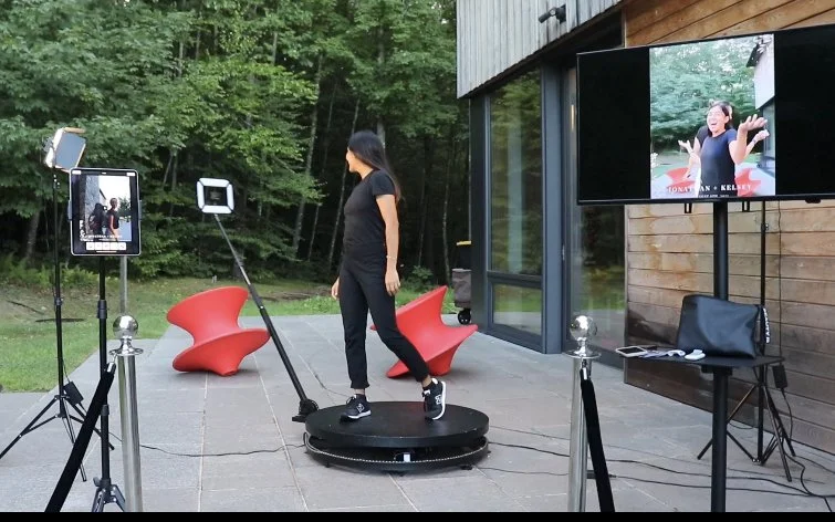 Woman dancing on a rotating platform during a virtual meeting or event, with screens showing her on video call, set outdoors with trees and modern seating nearby.